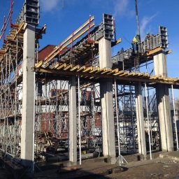 Matpol Budownictwo Sp.zo.o. - Wznoszenie konstrukcji żelbetowej budynku: szalunki kolumn i stropów, widoczni pracownicy w kaskach, rusztowania, w tle ceglany budynek i błękitne niebo.