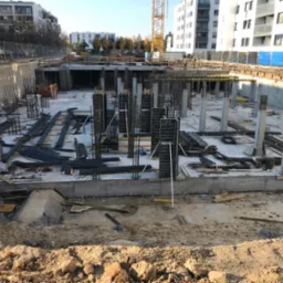 Wykop pod budynek wielorodzinny z widoczną konstrukcją zbrojeniową fundamentów i słupów, częściowo zaszalowaną, w tle budynki mieszkalne i żuraw budowlany.