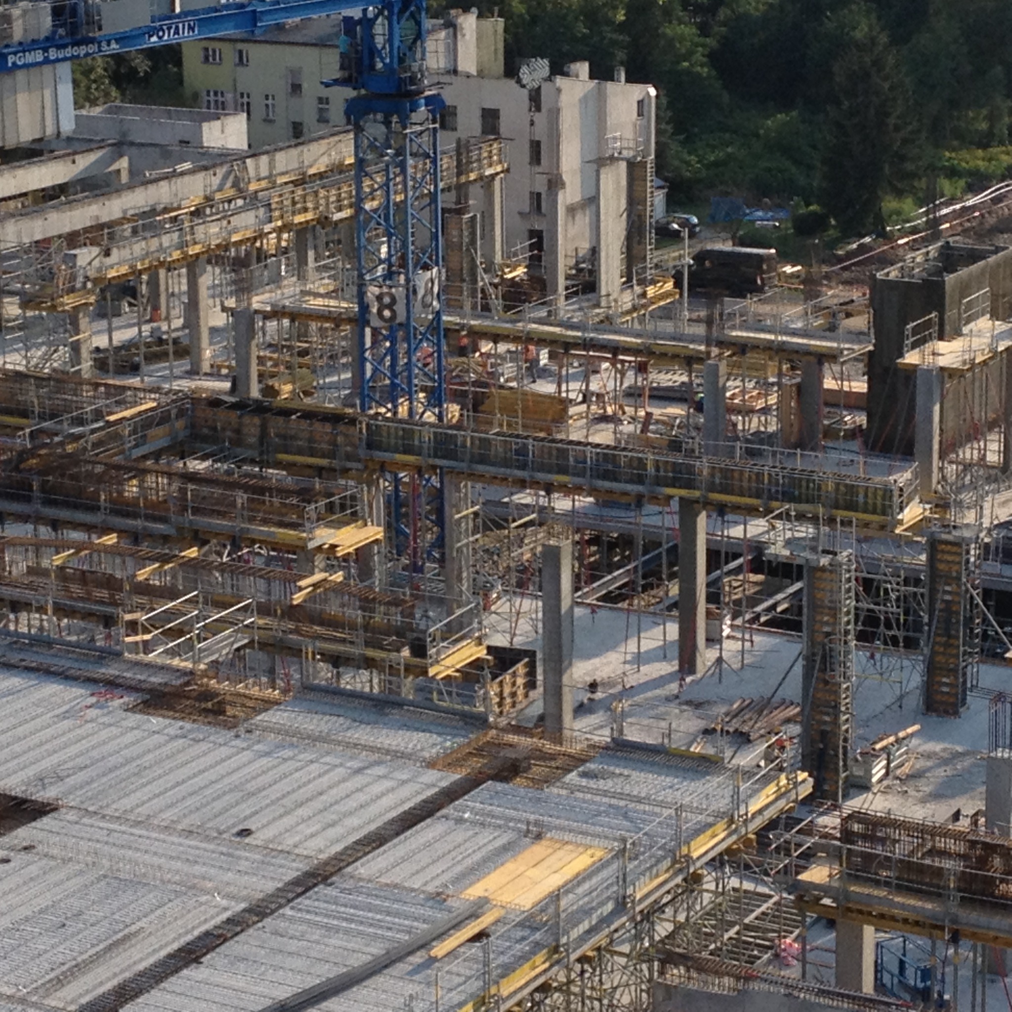 Wysoki kąt widzenia na plac budowy z widocznymi konstrukcjami szalunkowymi, zbrojeniem i elementami betonowymi w trakcie wznoszenia budynku, z dźwigiem budowlanym w tle.