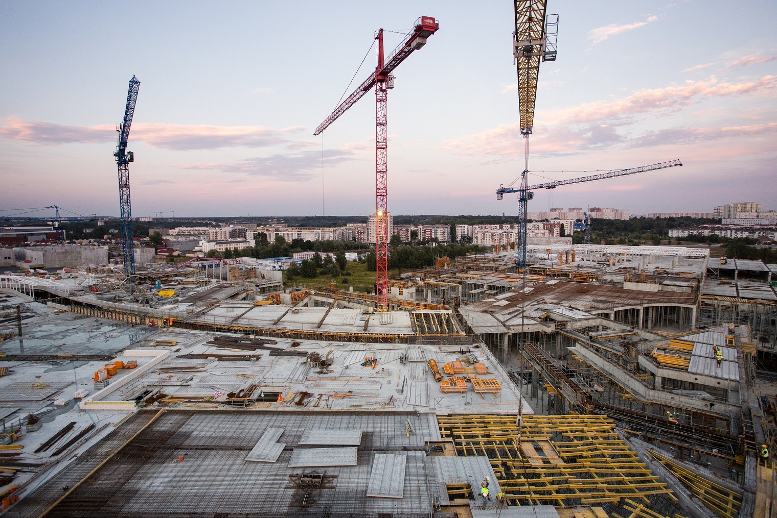 Rozległy plac budowy z widocznymi elementami szalunków, siatki zbrojeniowej, oraz czterema żurawiami wieżowymi na tle miejskiego krajobrazu o zmierzchu.