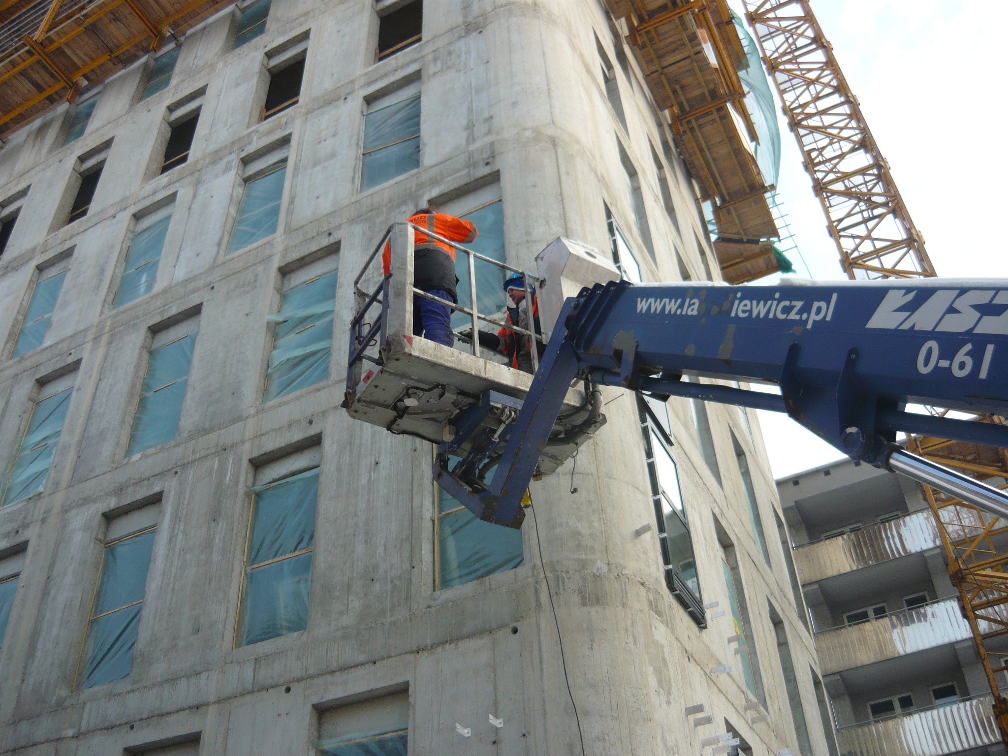 Dwóch robotników w pomarańczowej odzieży roboczej na podnośniku koszowym na tle surowego budynku z betonu, w tle widoczny żuraw budowlany.