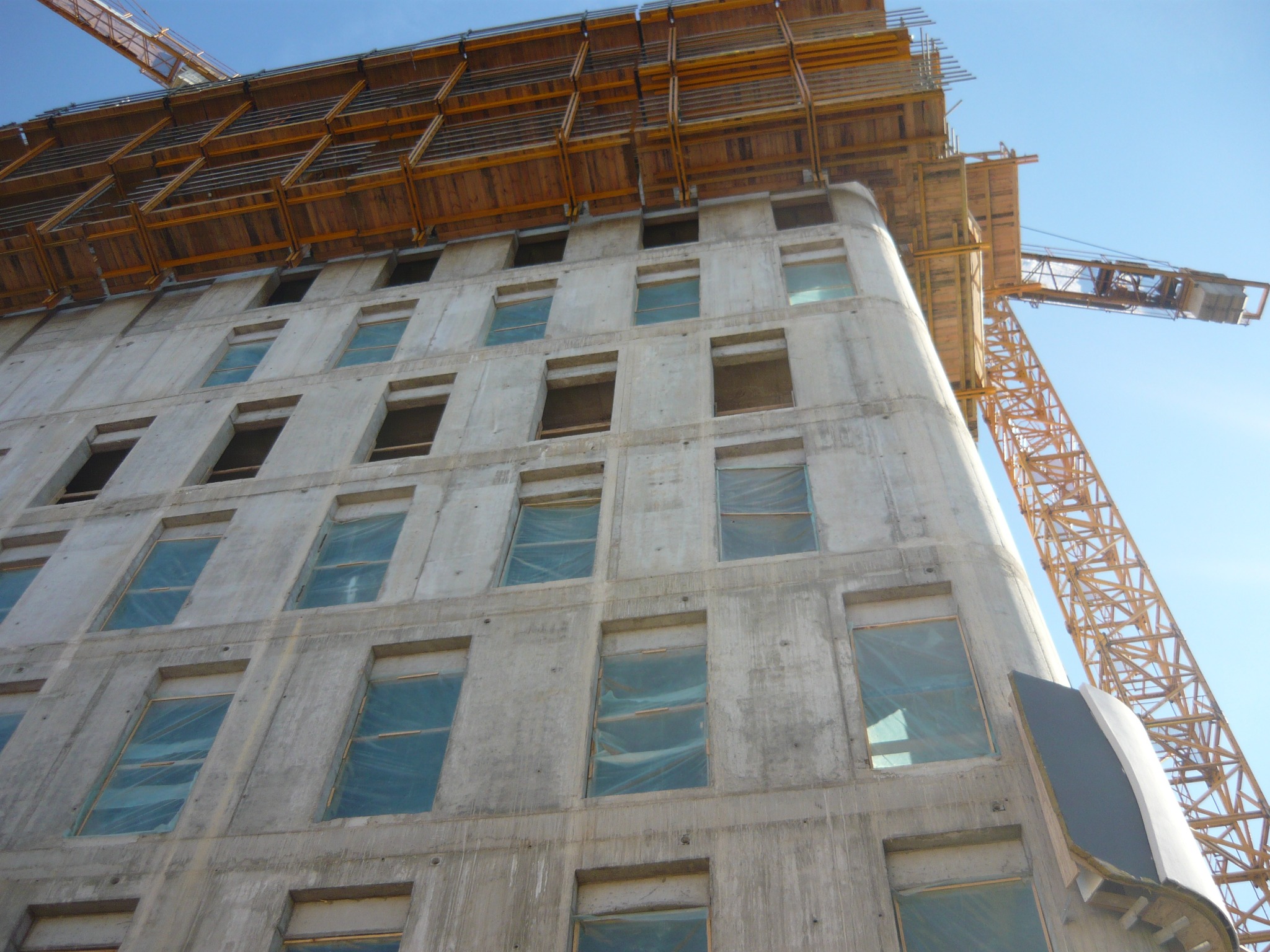 Wysoki budynek w trakcie budowy z betonu, z widocznymi otworami okiennymi zabezpieczonymi folią, otoczony rusztowaniami i dźwigiem budowlanym na tle błękitnego nieba.