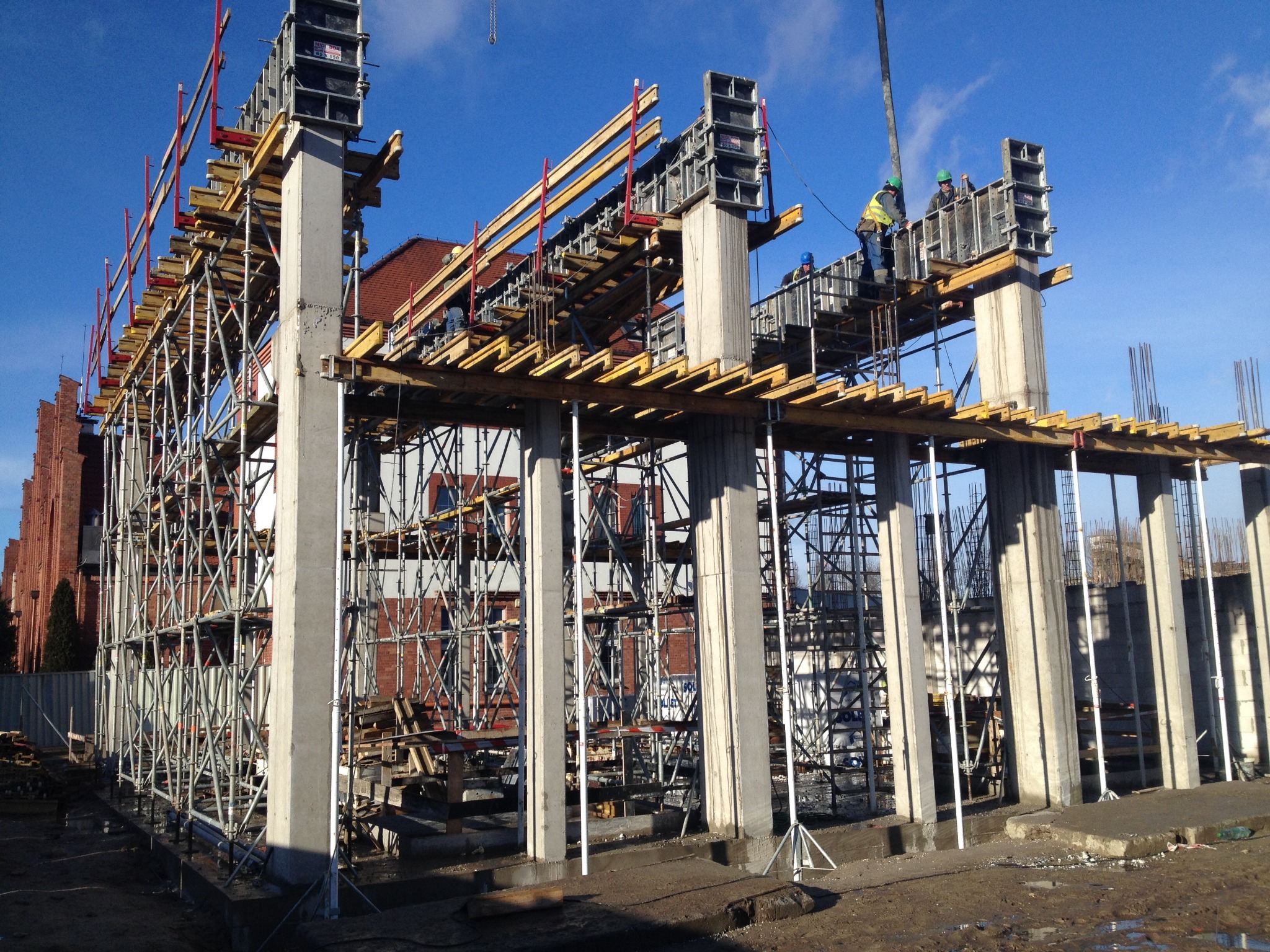Wznoszenie konstrukcji żelbetowej budynku: szalunki kolumn i stropów, widoczni pracownicy w kaskach, rusztowania, w tle ceglany budynek i błękitne niebo.