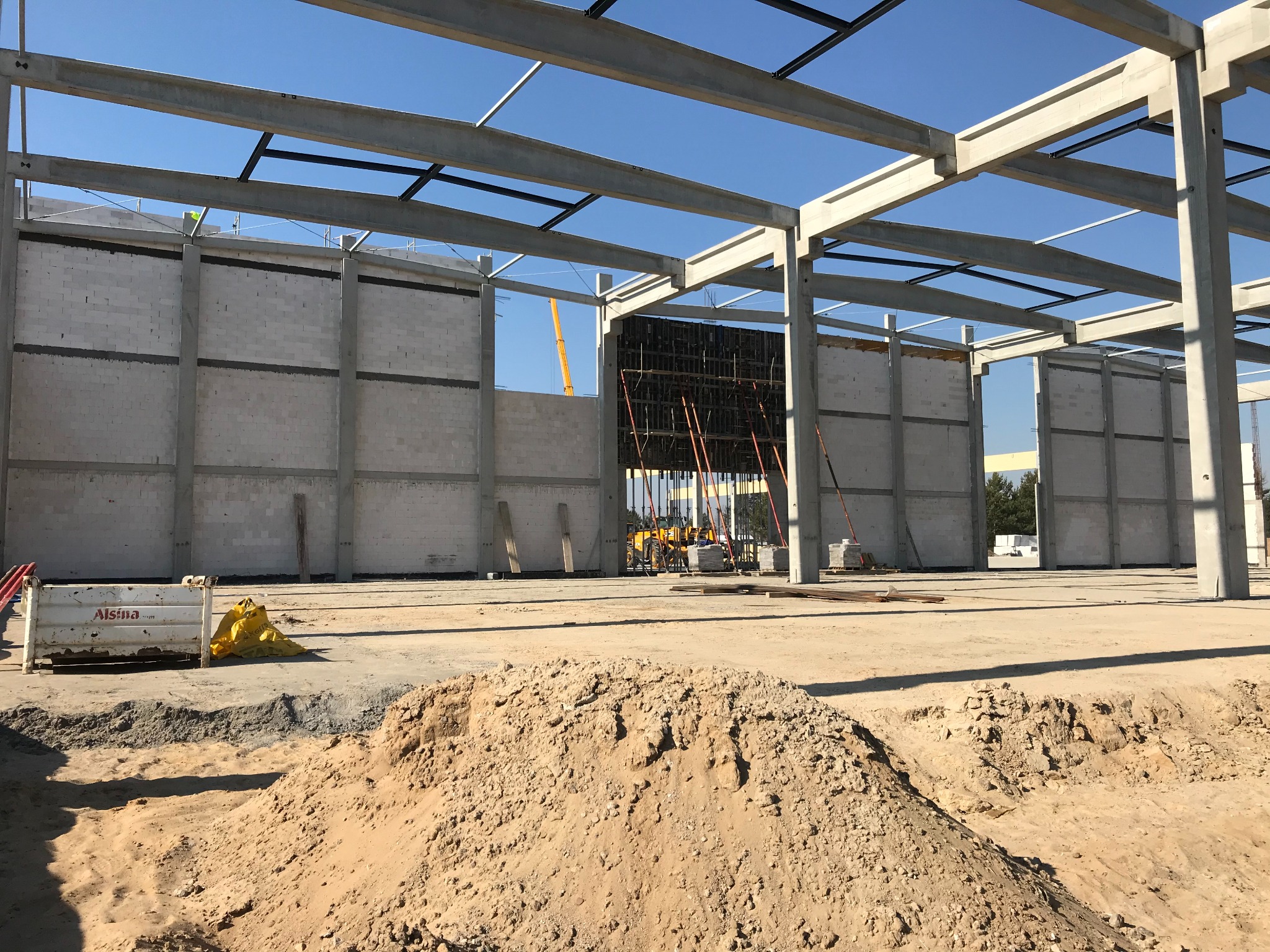Budowa hali żelbetowej w trakcie wznoszenia, widoczne szkielet konstrukcji, ściany z bloczków betonowych, żółty żuraw w tle i hałda piasku na pierwszym planie.