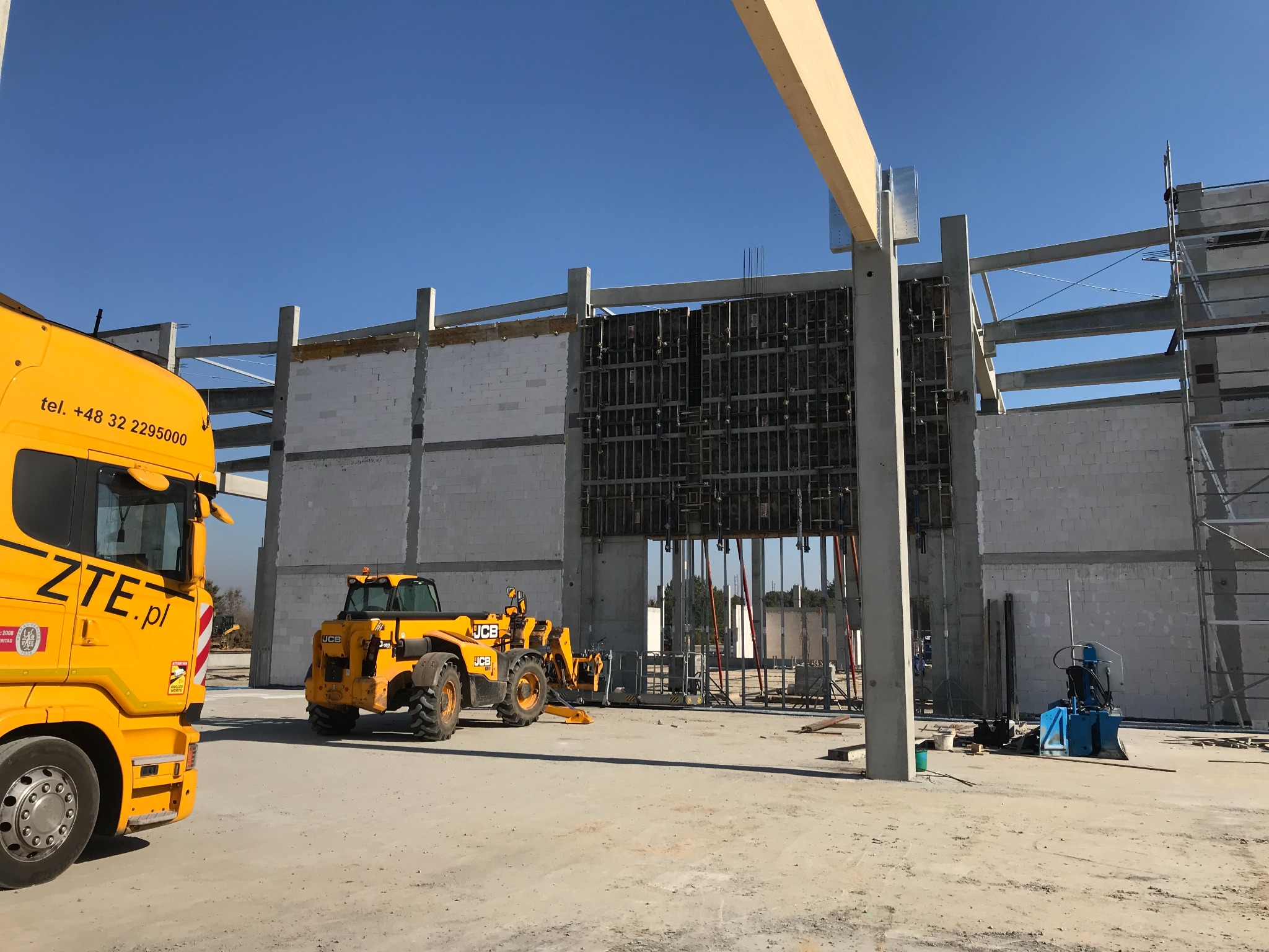 Wznoszenie budynku z betonu komórkowego z widocznymi szalunkami, żółty teleskopowy wózek widłowy JCB i ciężarówka z logo ZTE.pl na placu budowy w słoneczny dzień.