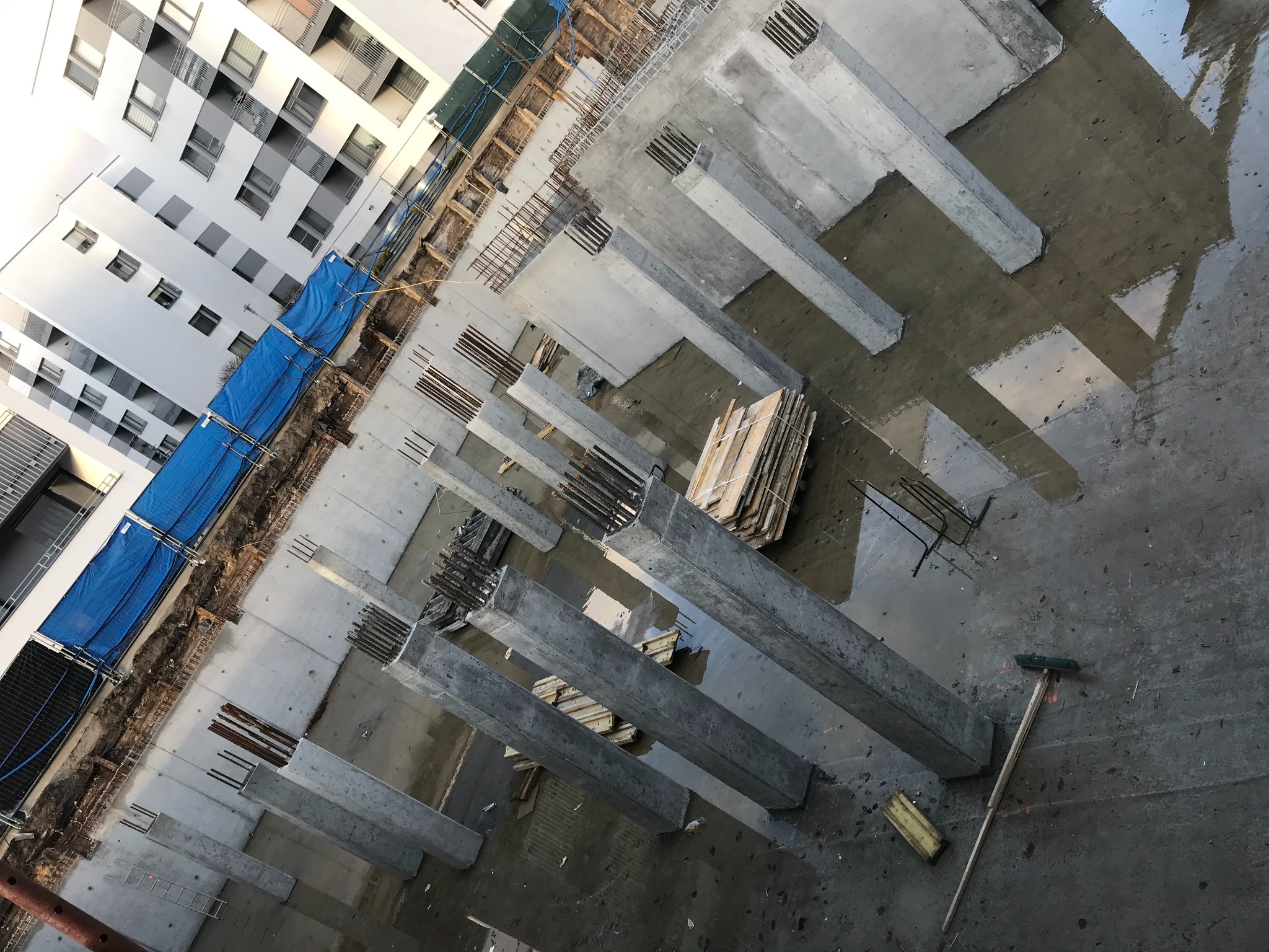 Wysoki kąt widzenia na plac budowy z prefabrykowanymi, betonowymi słupami z wystającą zbrojeniem, częściowo zalany teren, w tle budynek mieszkalny z niebieską siatką ochronną.