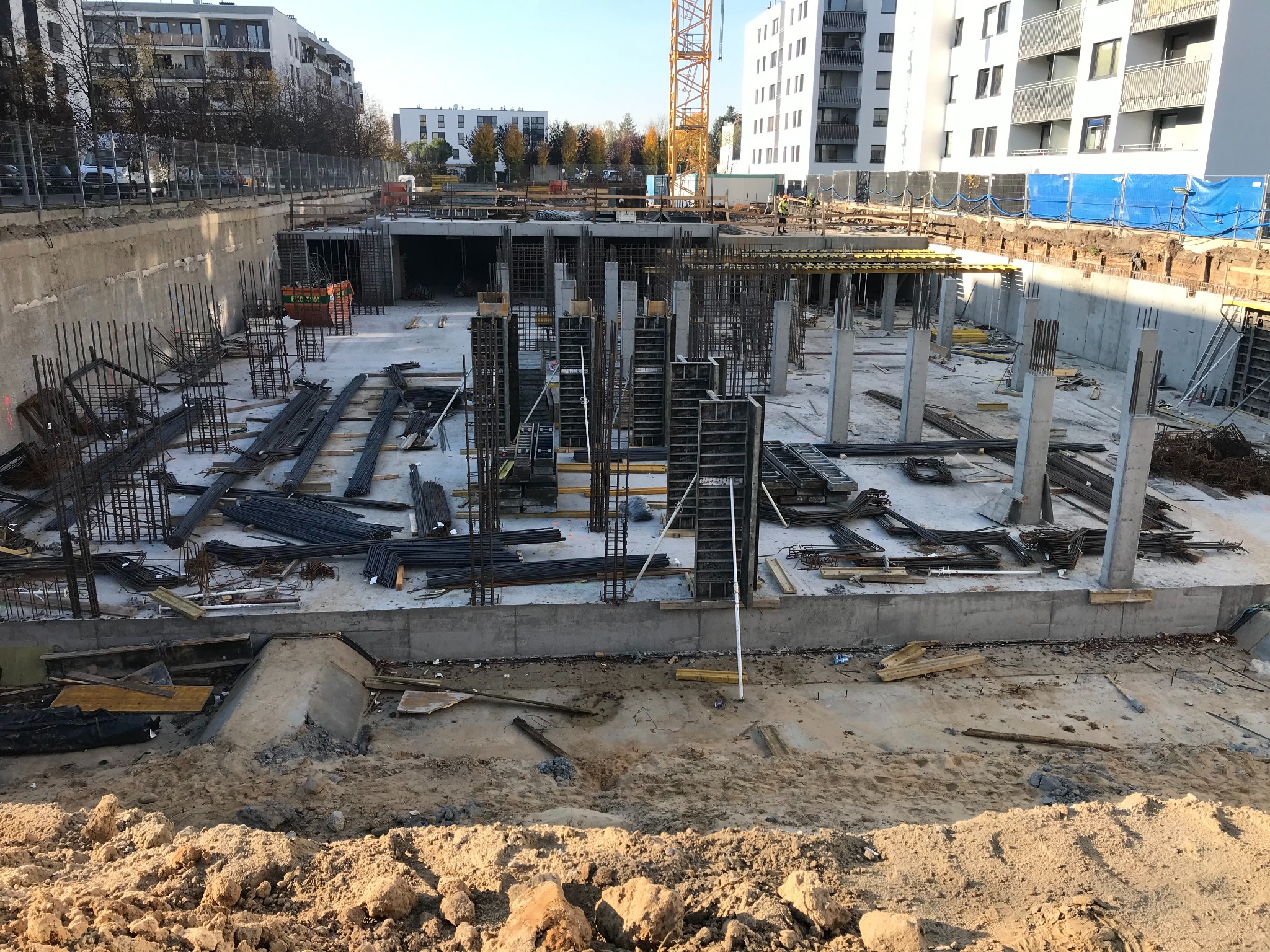 Wykop pod budynek wielorodzinny z widoczną konstrukcją zbrojeniową fundamentów i słupów, częściowo zaszalowaną, w tle budynki mieszkalne i żuraw budowlany.