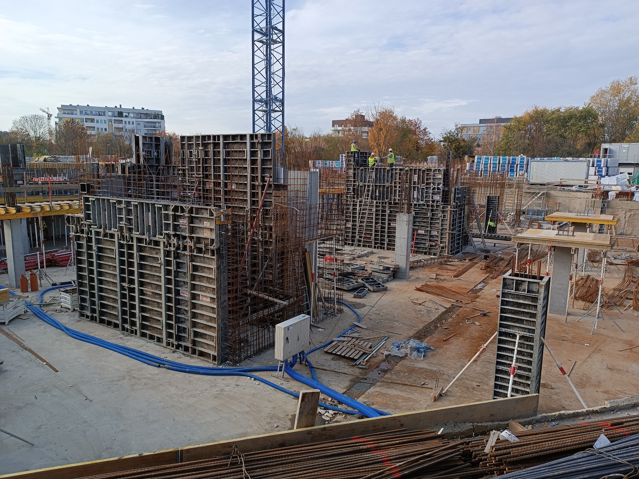 Wykonywanie szalunków na budowie z widocznymi pracownikami w odblaskowych kamizelkach i niebieskimi wężami doprowadzającymi media.