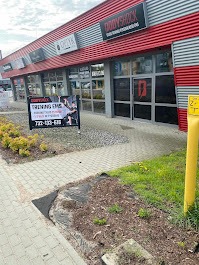 Zewnętrzny widok budynku z szyldem 'BODYSHOCK TRAINING EMS' i tablicą reklamową z informacjami o treningach EMS oraz numerem telefonu, widoczne elementy krajobrazu miejskiego i ścieżka z kostki...