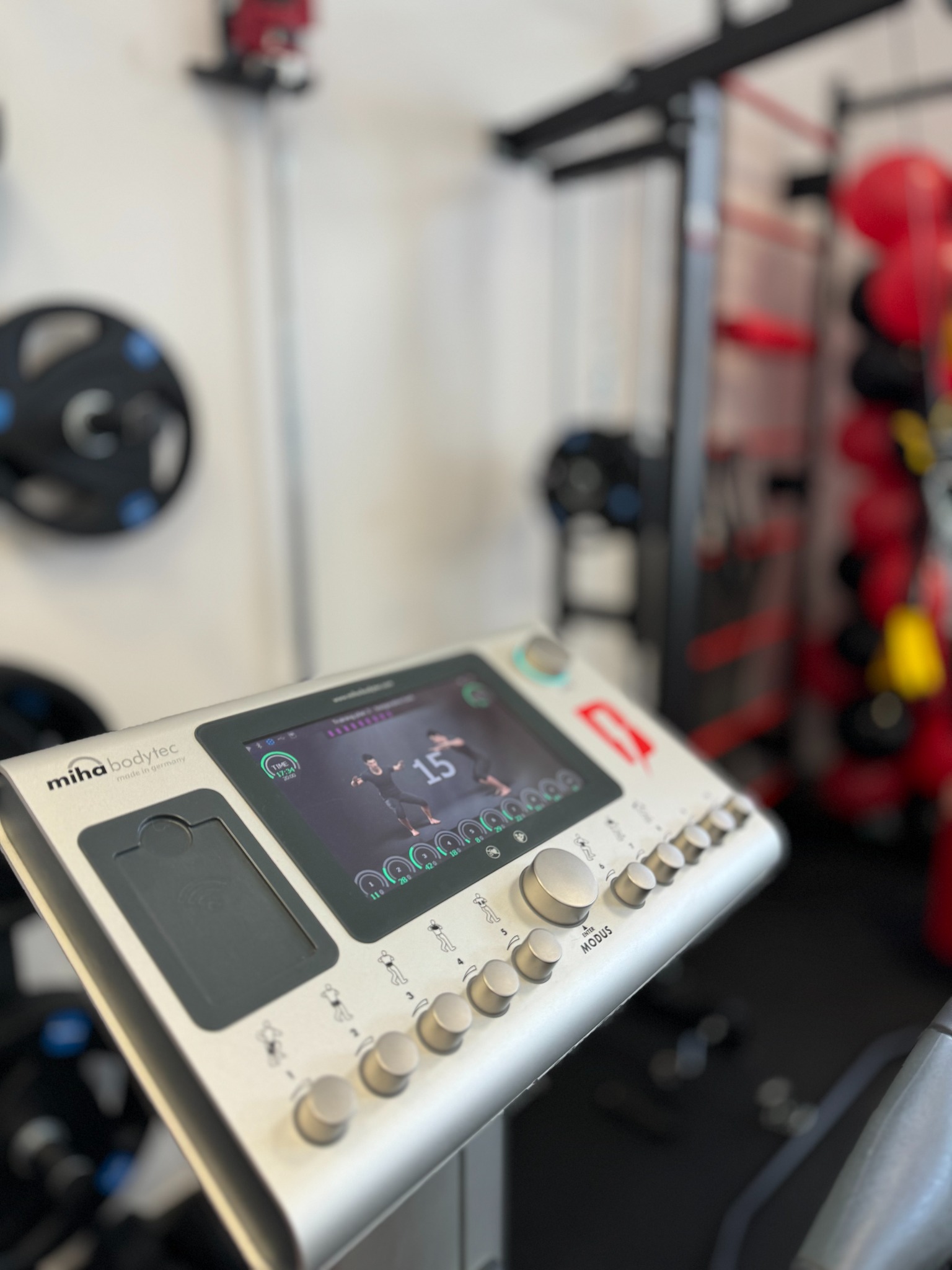 Urządzenie Miha Bodytec do elektrostymulacji mięśni, ekran pokazuje ćwiczenia, w tle sprzęt do ćwiczeń i piłki.