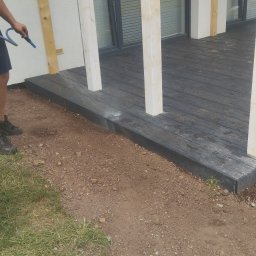 Firma budowlana trójca - Czyszczenie wodą drewnianego tarasu z jasnymi słupami, widoczny fragment trawnika i ziemi.