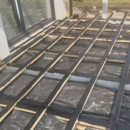 Firma budowlana trójca - Przygotowanie podłoża pod taras: Czarne folie izolacyjne i drewniane legary pomalowane na czarno, ułożone na białym podłożu. W tle widoczny fragment budynku z oknem i mężczyzna w pomarańczowych...