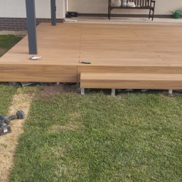 Firma budowlana trójca - Nowo wybudowany taras z kompozytowych desek w ogrodzie, widoczne ślady po pracy, wkrętarka i nóż monterski leżą na trawie.