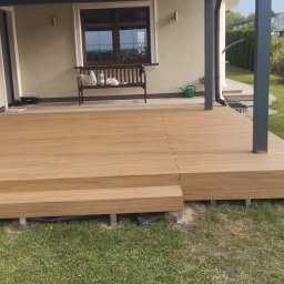 Firma budowlana trójca - Nowoczesny taras z desek kompozytowych w kolorze drewna, z dwoma stopniami, widok na dom z oknem i ławką oraz fragment ogrodu.