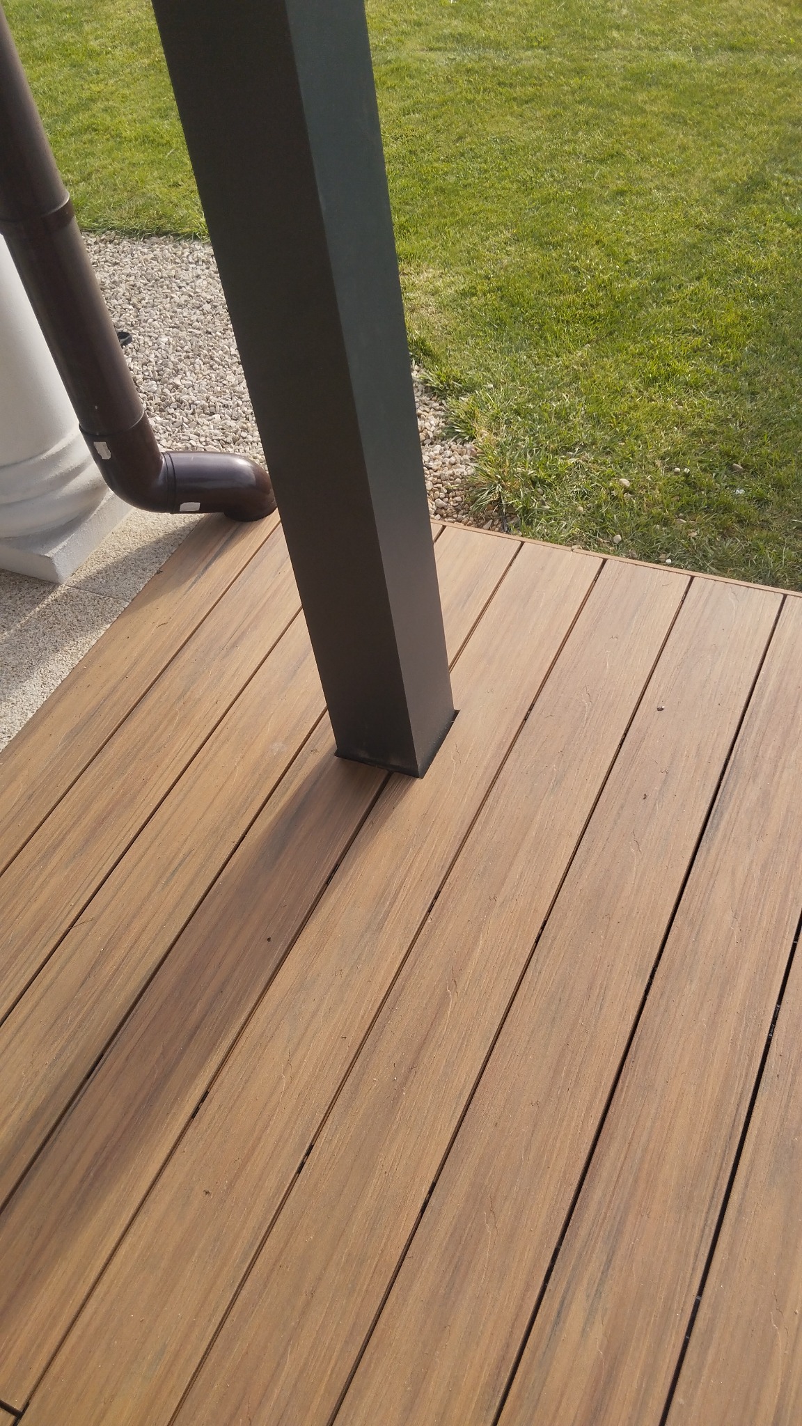 Drewniany taras z desek kompozytowych w odcieniach brązu, z czarną kwadratową kolumną podtrzymującą zadaszenie, widoczny fragment rynny i trawnika w tle.