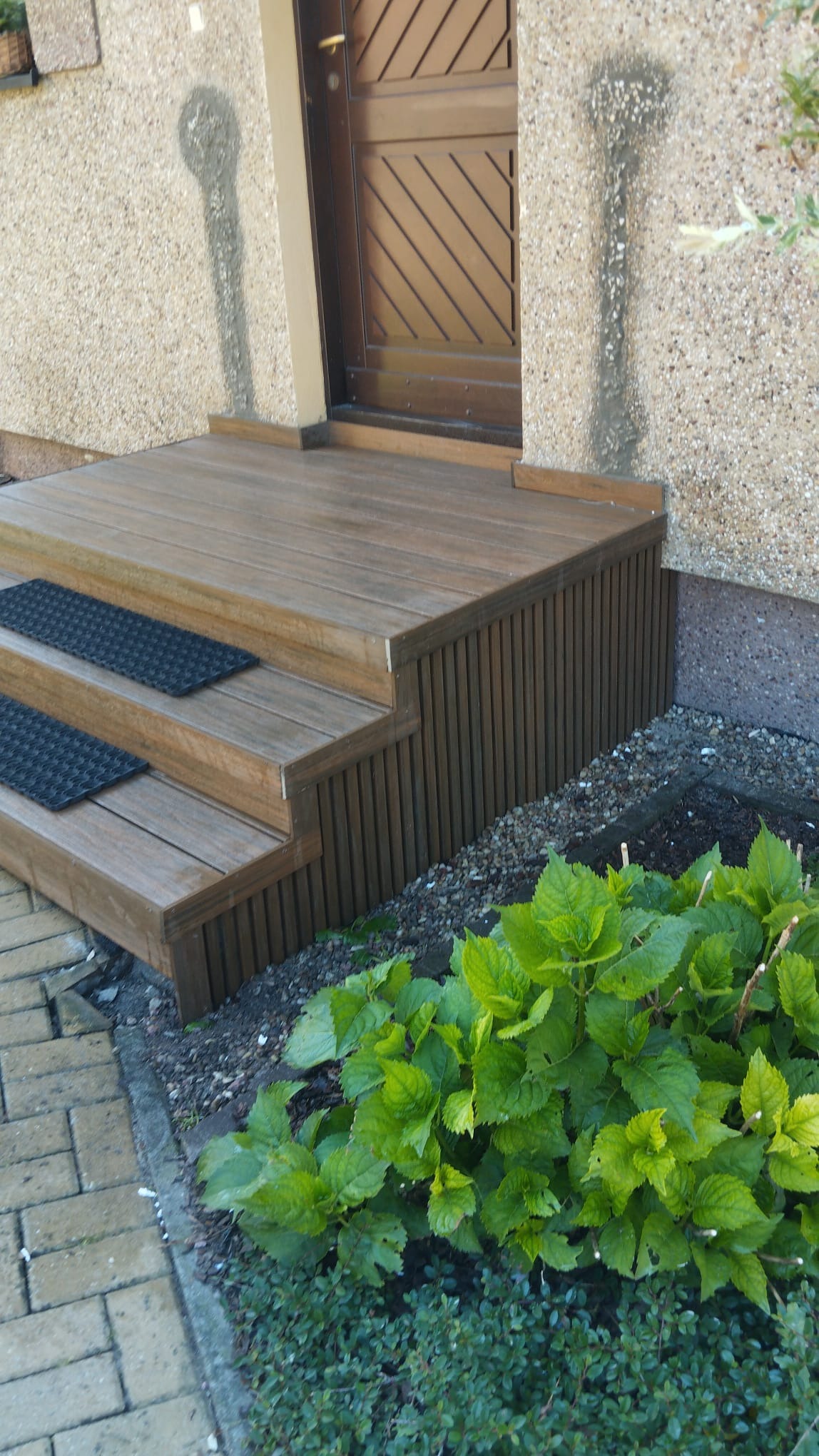 Drewniane schody wejściowe z dwoma czarnymi wycieraczkami prowadzące do brązowych drzwi, obok kwitnące krzewy hortensji.