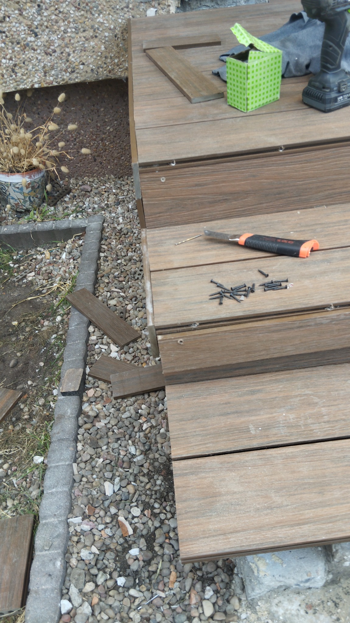 Montaż tarasu kompozytowego: widoczne deski, wkręty, nóż, wiertarka akumulatorowa, pudełko po wkrętach i rękawice robocze na tle kamienistego podłoża.