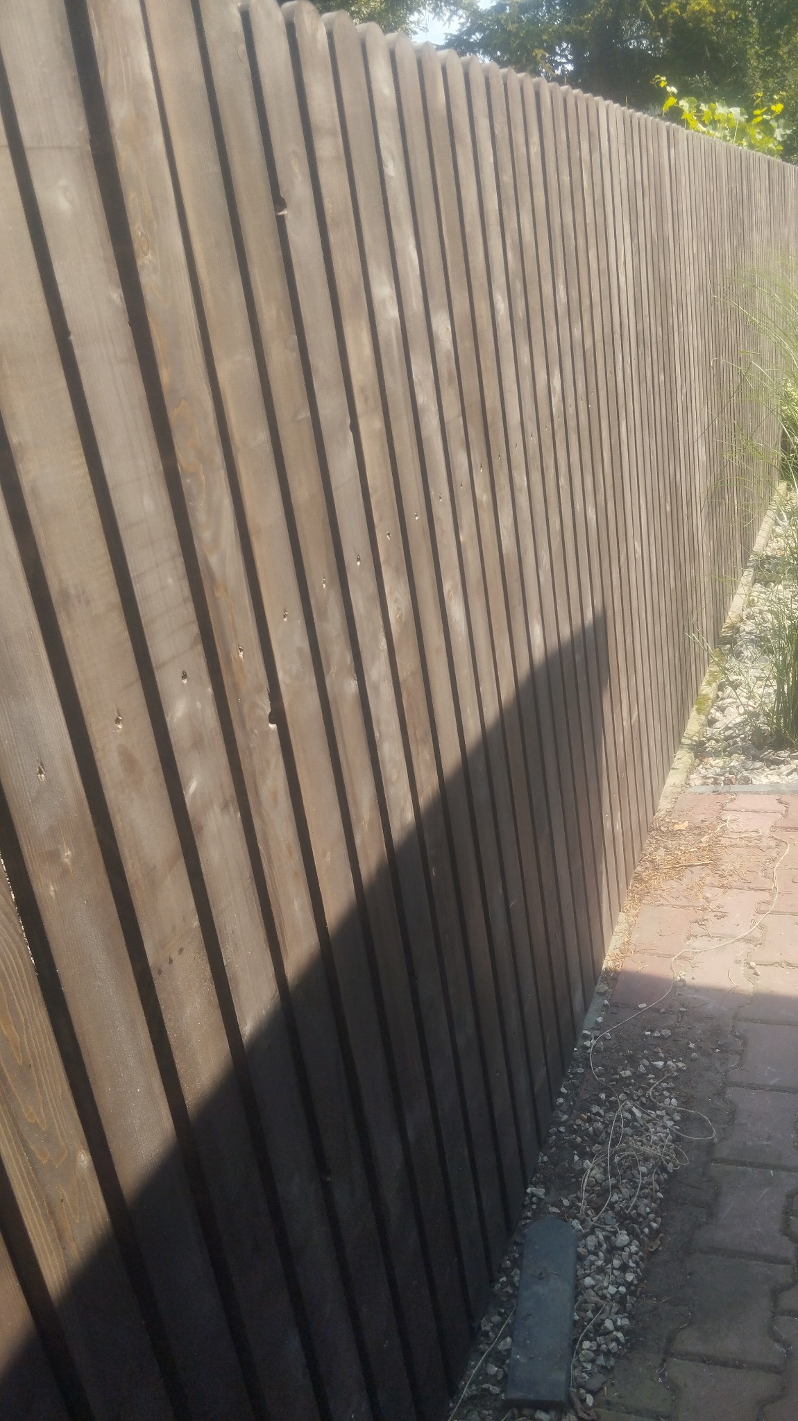 Drewniany płot z pionowych desek, widoczny cień, obok ścieżka z kostki brukowej i kamyczki.