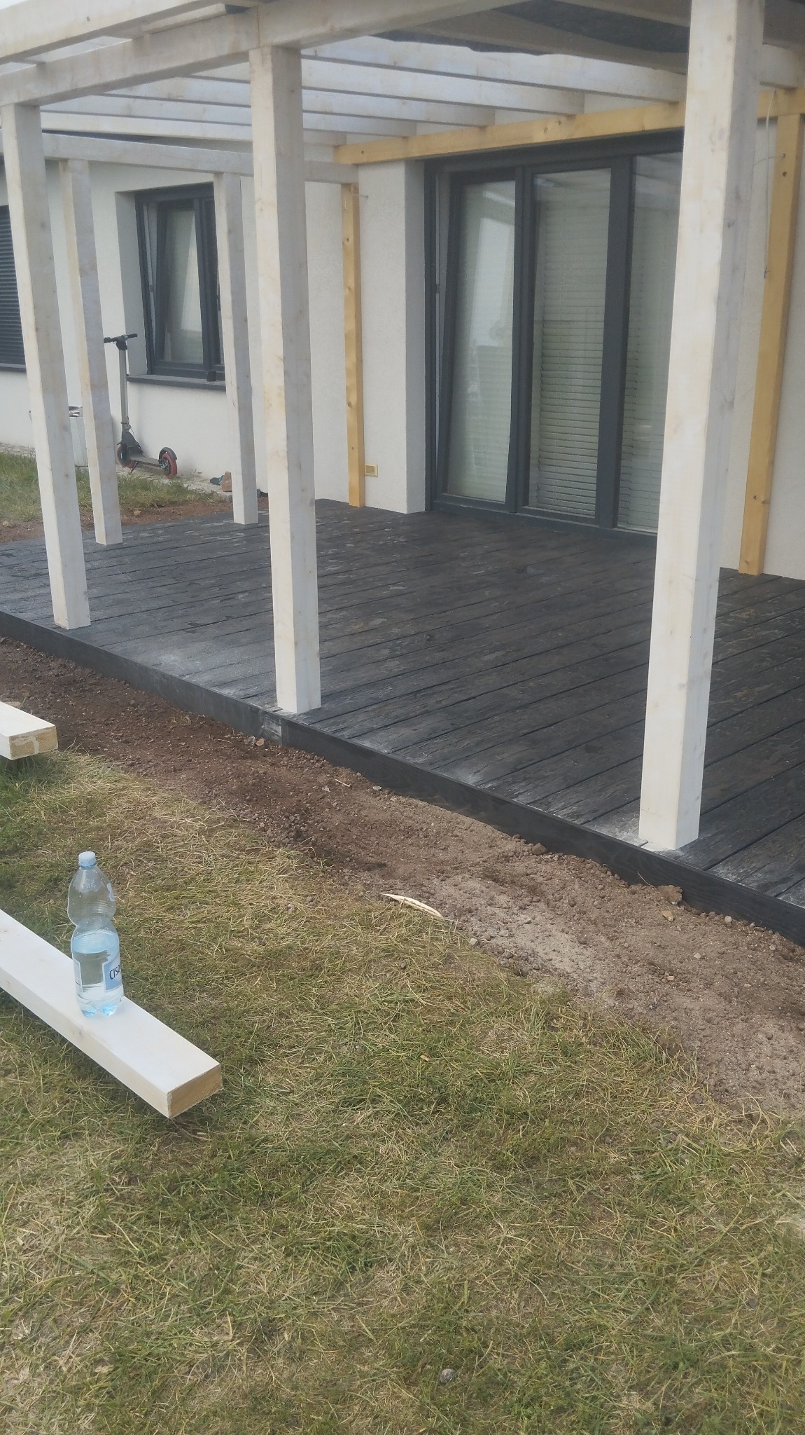 Montaż drewnianego tarasu z pergolą przy nowoczesnym domu, widoczna butelka wody na desce, trawa i częściowo wykończona rabata.