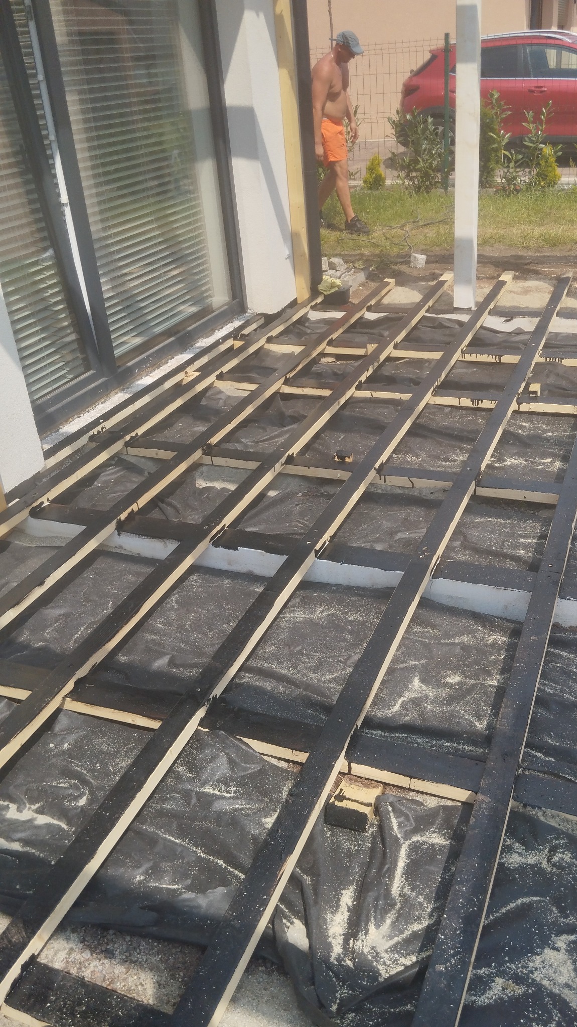 Przygotowanie podłoża pod taras: Czarne folie izolacyjne i drewniane legary pomalowane na czarno, ułożone na białym podłożu. W tle widoczny fragment budynku z oknem i mężczyzna w pomarańczowych...