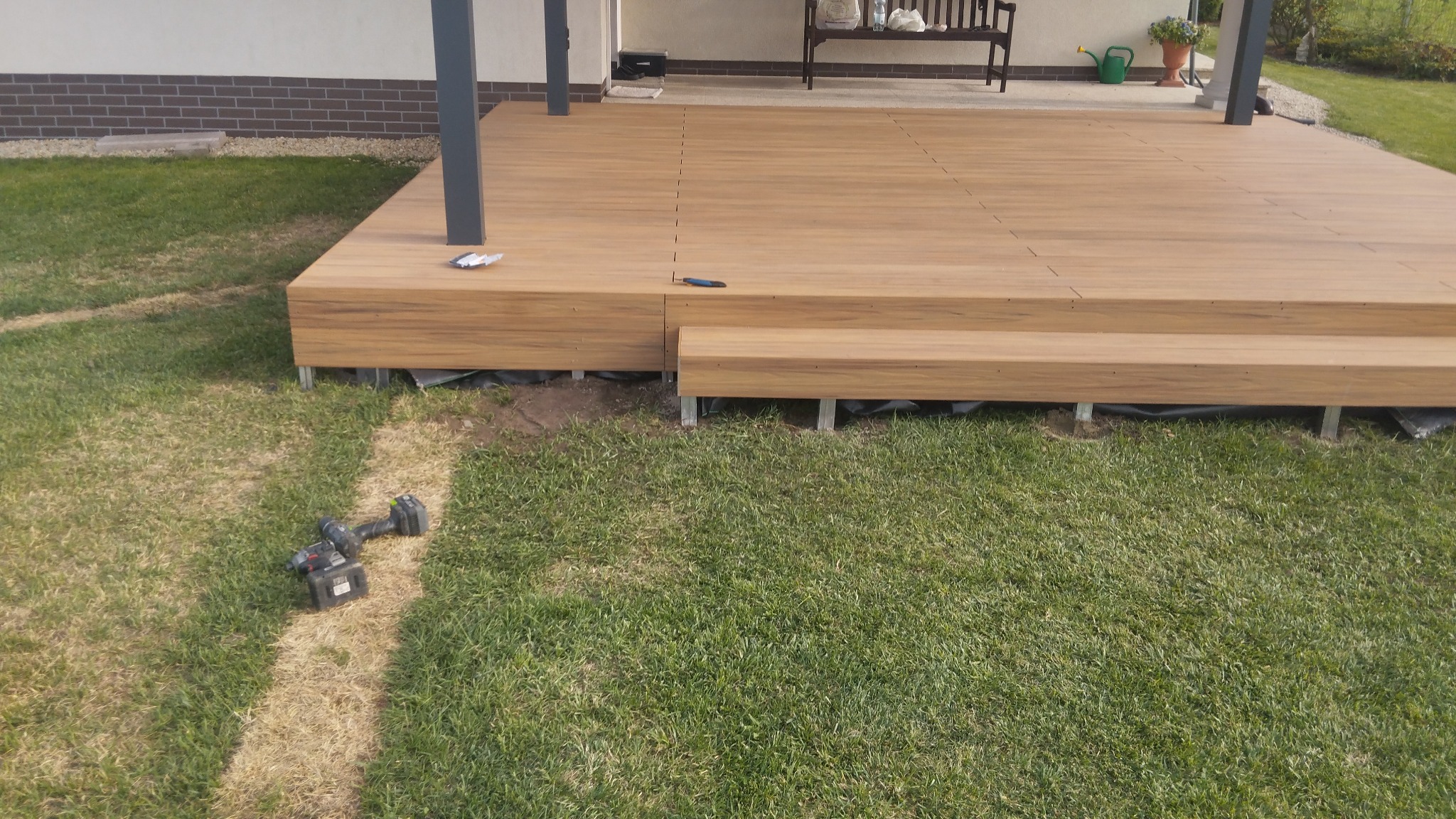 Nowo wybudowany taras z kompozytowych desek w ogrodzie, widoczne ślady po pracy, wkrętarka i nóż monterski leżą na trawie.