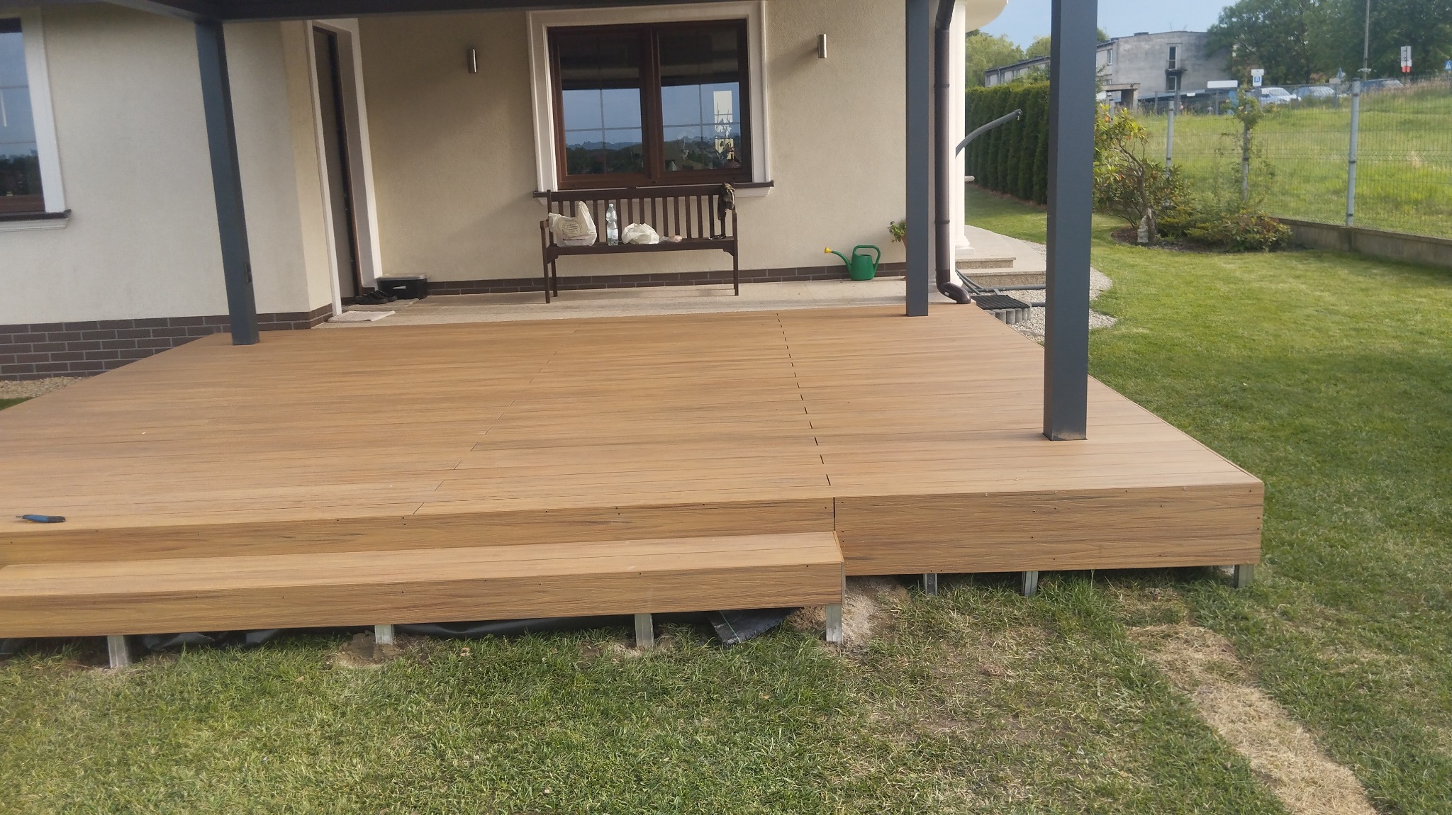 Nowoczesny taras z desek kompozytowych w kolorze drewna, z dwoma stopniami, widok na dom z oknem i ławką oraz fragment ogrodu.