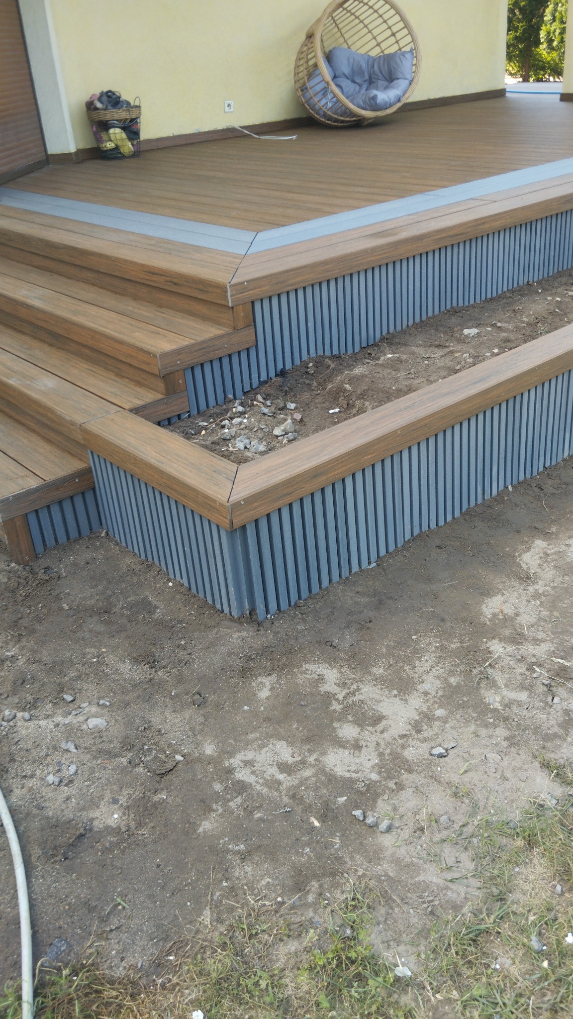 Drewniany taras z kompozytu z schodami i rabatą, widok z dołu, częściowo ukończony, z ziemią wokół.