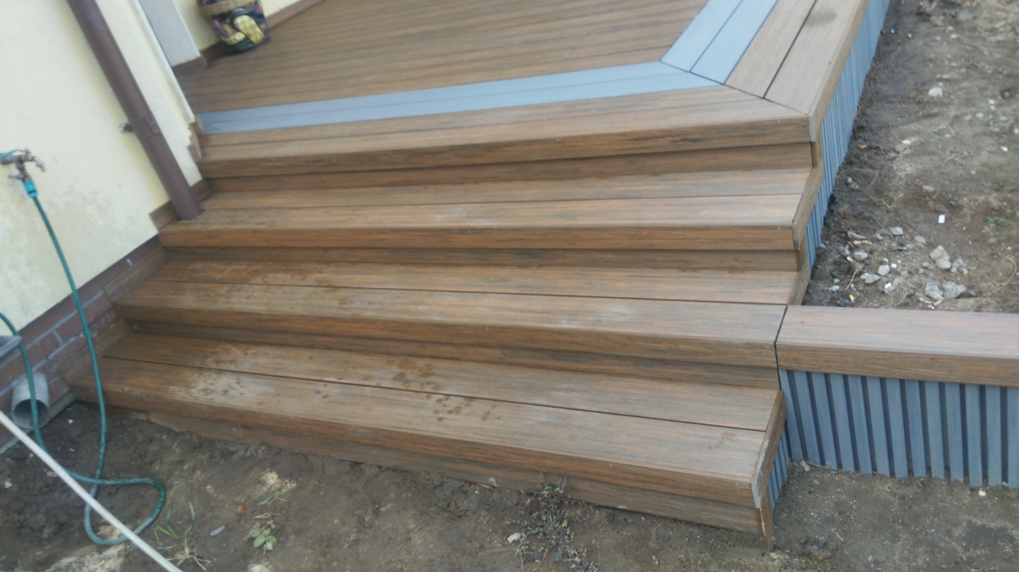 Schody zewnętrzne z kompozytu drewna prowadzące do tarasu, z widocznym fragmentem elewacji budynku i ziemią wokół konstrukcji.