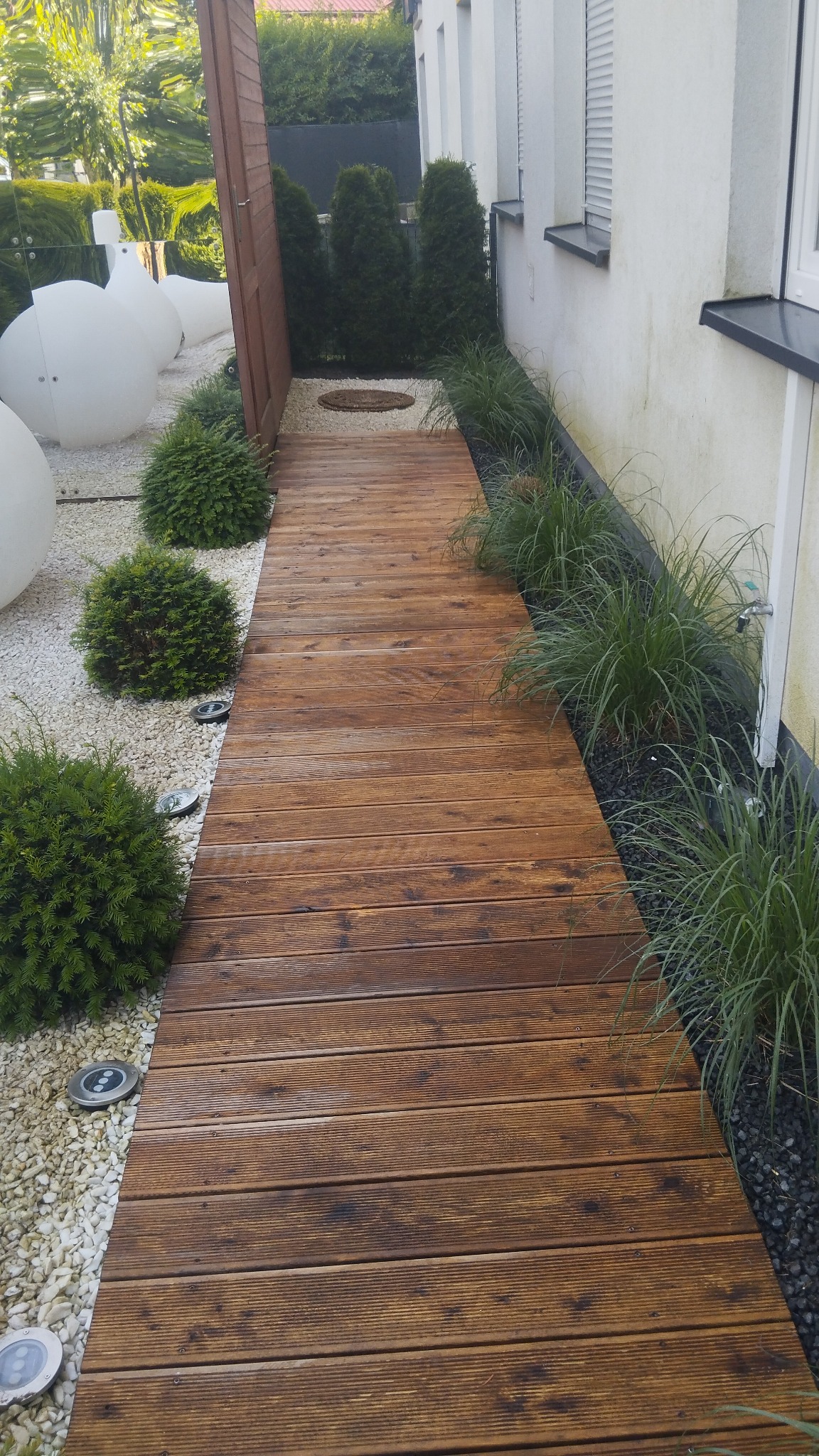 Drewniana ścieżka z desek prowadząca przez ogród, oświetlona lampami gruntowymi, z białym i czarnym żwirem oraz roślinami ozdobnymi po bokach, przylegająca do ściany budynku.