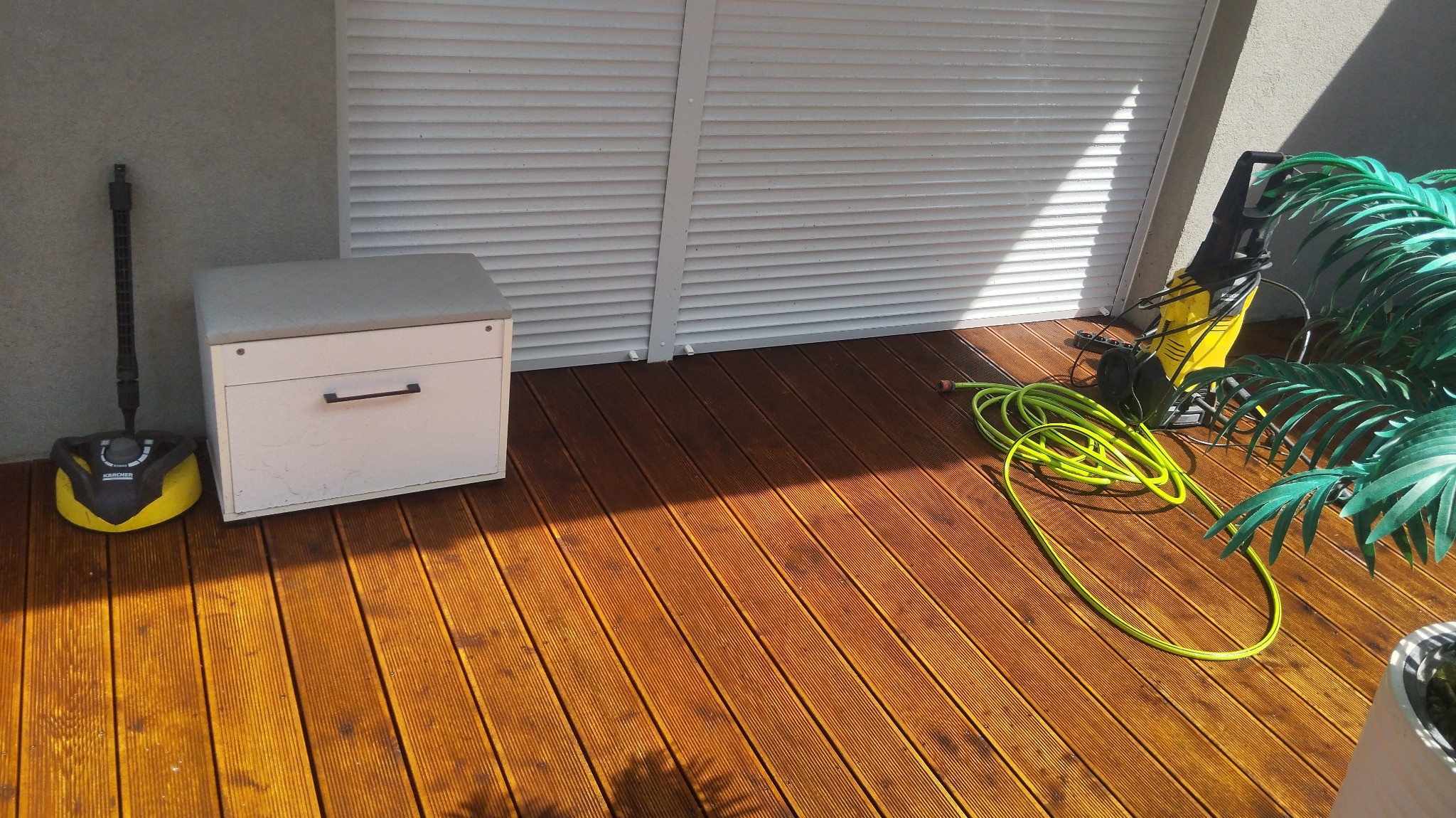 Wyczyszczony fragment drewnianego tarasu z widoczną deską podłogową po prawej stronie, obok żółtej myjki ciśnieniowej z zielonym wężem, a po lewej stronie skrzynia z siedziskiem i żółta szczotka...