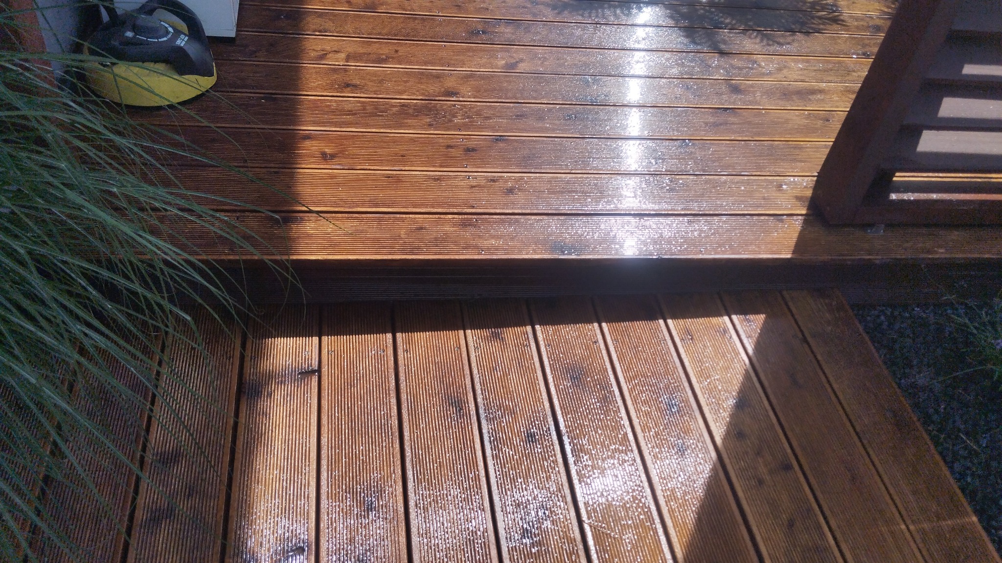 Świeżo umyta drewniana podłoga tarasu z widocznymi kroplami wody, z fragmentem rośliny w lewym górnym rogu i żółtym urządzeniem czyszczącym w tle.