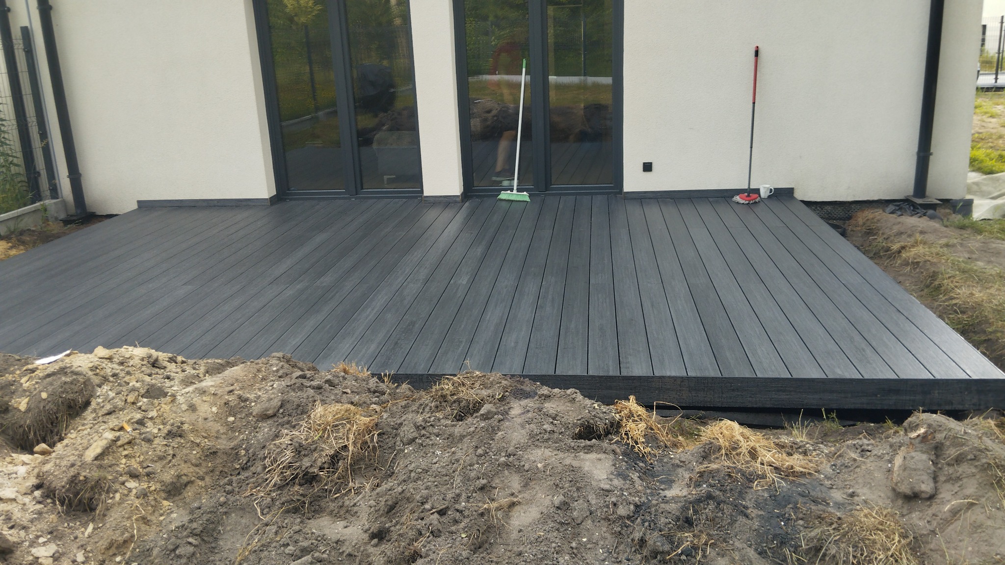 Nowoczesny taras z kompozytu w kolorze grafitowym, widok z perspektywy ziemi, z widocznym wykopem i stertą ziemi w pierwszym planie, przy ścianie domu z dużymi, przeszklonymi drzwiami.