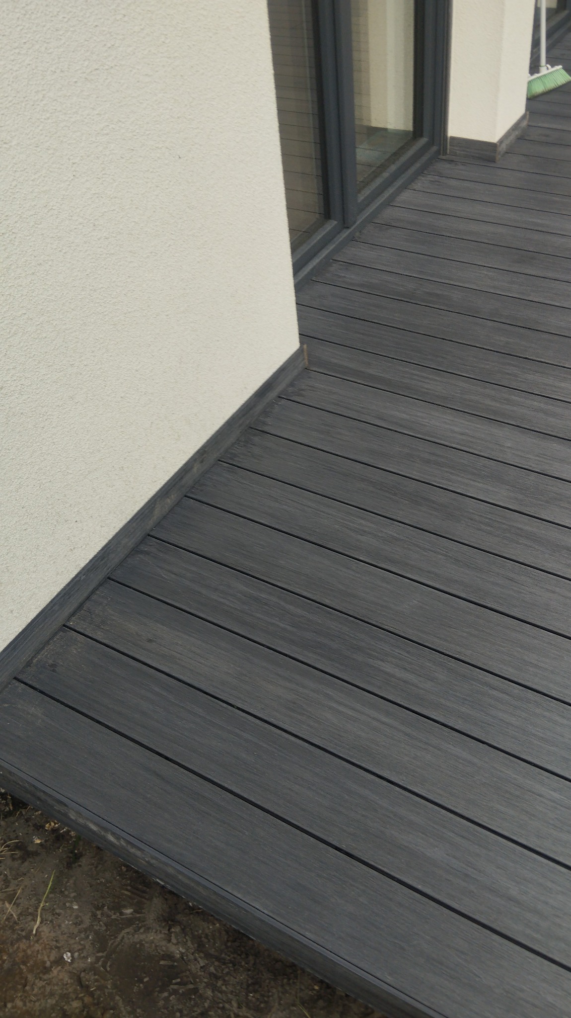 Nowoczesny taras z desek kompozytowych w odcieniach szarości, przylegający do ściany budynku z widocznym oknem i fragmentem elewacji o fakturze baranka.