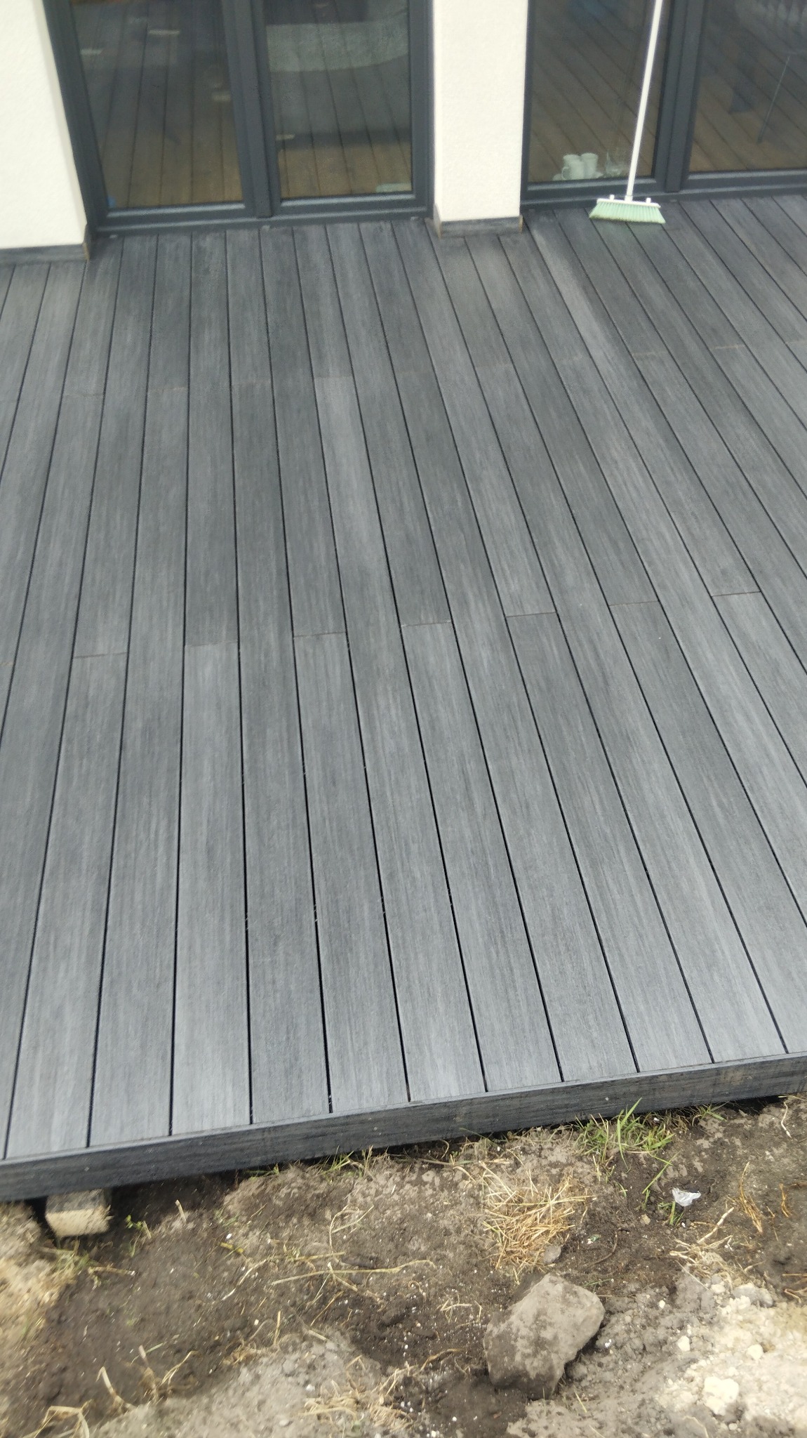 Nowo wybudowany taras z desek kompozytowych w odcieniach szarości, widok z góry, z fragmentem niezabudowanego terenu z ziemią i trawą przy krawędzi tarasu, częściowo przysłonięty miotłą opartą o okno.