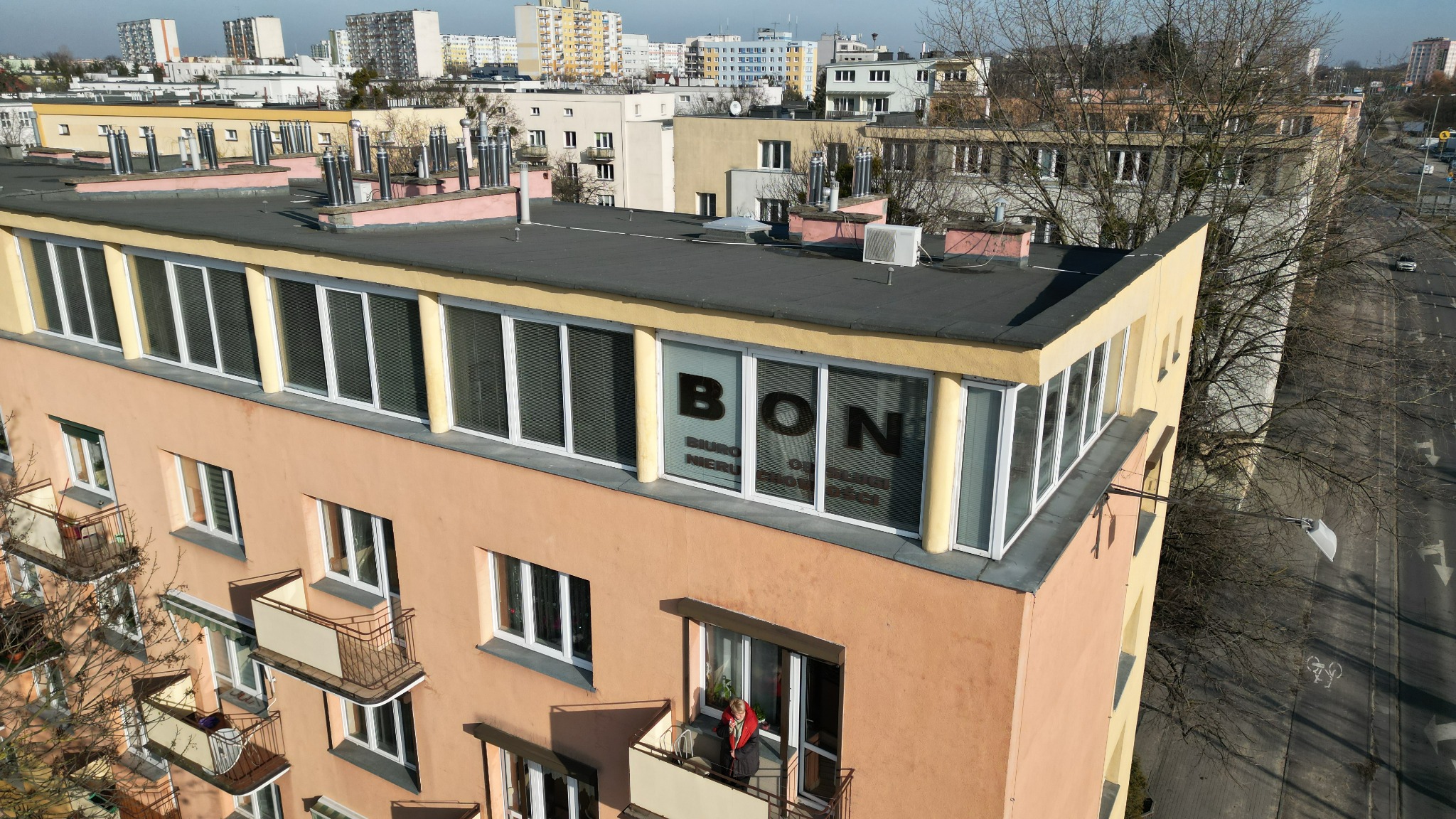 Budynek biura nieruchomości 'BON' z widokiem z góry na balkony i ulicę, z widocznymi drzewami bez liści.