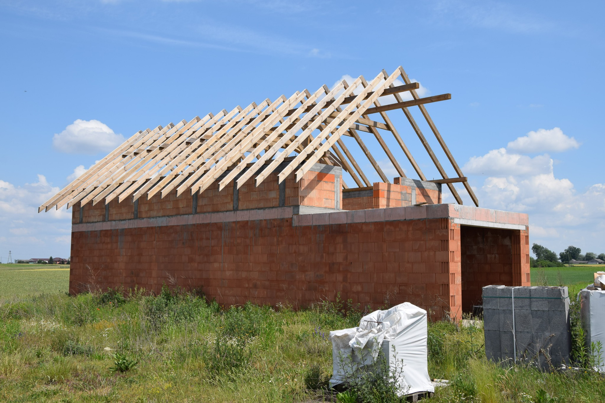 Surowy stan budynku z czerwonej cegły z drewnianą konstrukcją dachu, otoczony wysoką trawą i paletami materiałów budowlanych.