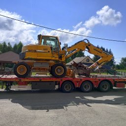 Raptor-Trans Erik Sleziona - Żółta koparka kołowa Liebherr 309 na czerwonej niskopodwoziowej platformie transportowej, zabezpieczona łańcuchami, na tle domów i błękitnego nieba z chmurami.