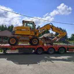 Żółta koparka kołowa Liebherr 309 na czerwonej niskopodwoziowej platformie transportowej, zabezpieczona łańcuchami, na tle domów i błękitnego nieba z chmurami.