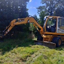 Raptor-Trans Erik Sleziona - Żółta koparka kołowa marki Liebherr stoi na trawiastym terenie z drzewami w tle, z widoczną łyżką i lemieszem. Kabina operatora jest otwarta, a obok stoi metalowa beczka.