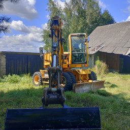 Raptor-Trans Erik Sleziona - Żółta koparka kołowa stoi na trawiastym terenie, z czarną łyżką skierowaną w stronę obiektywu, na tle budynku gospodarczego i błękitnego nieba z chmurami.