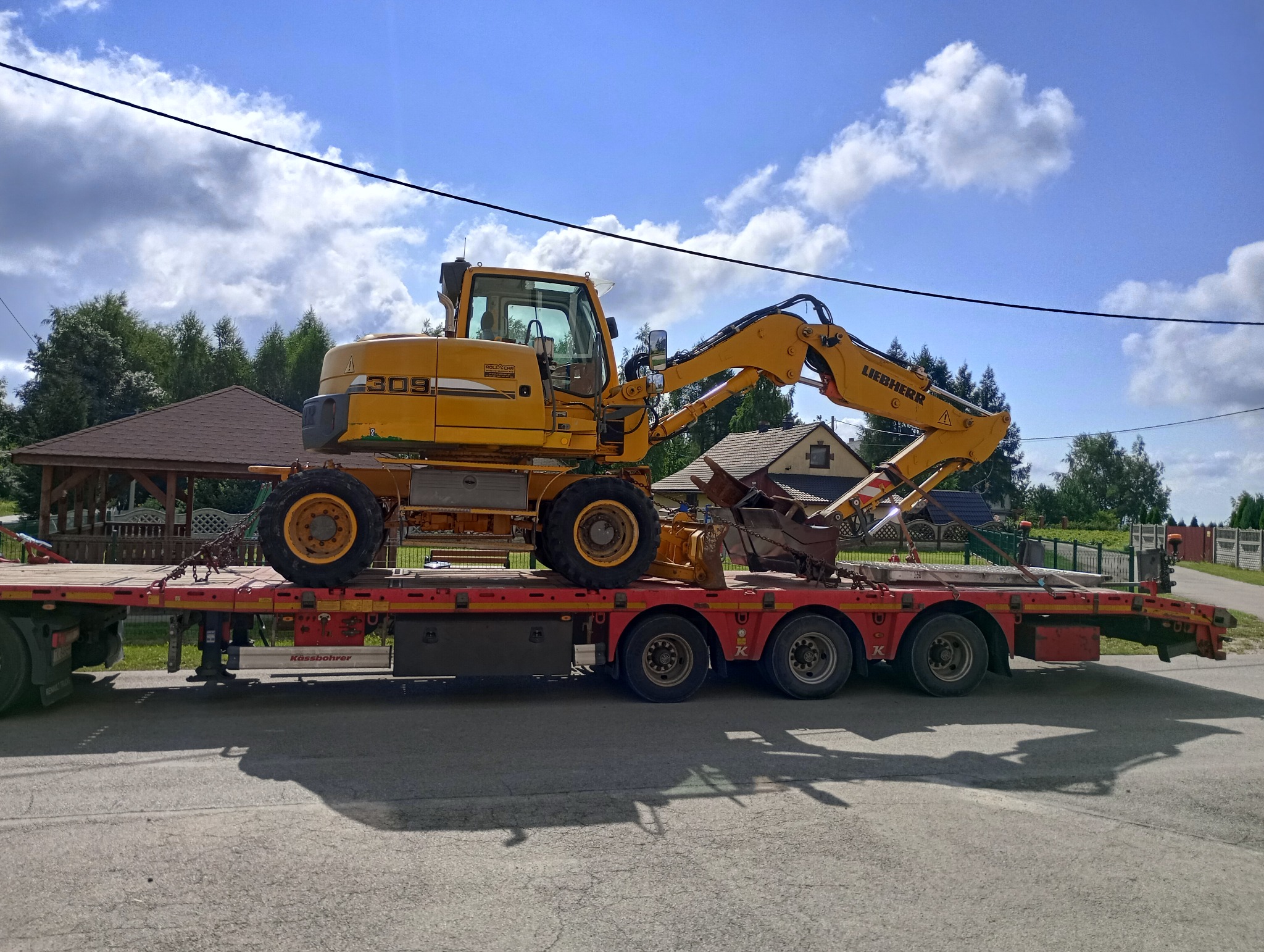 Żółta koparka kołowa Liebherr 309 na czerwonej niskopodwoziowej platformie transportowej, zabezpieczona łańcuchami, na tle domów i błękitnego nieba z chmurami.