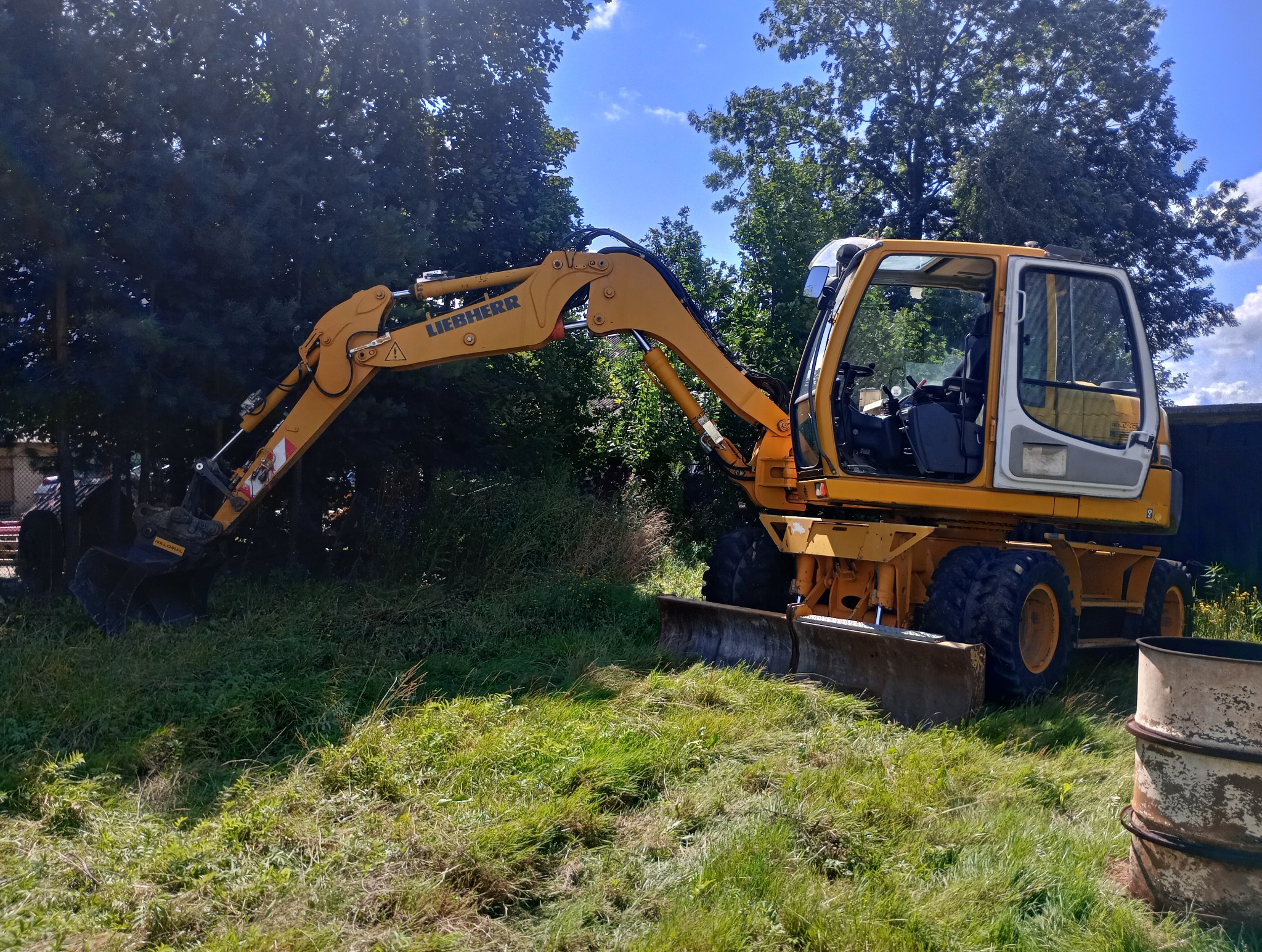 Żółta koparka kołowa marki Liebherr stoi na trawiastym terenie z drzewami w tle, z widoczną łyżką i lemieszem. Kabina operatora jest otwarta, a obok stoi metalowa beczka.