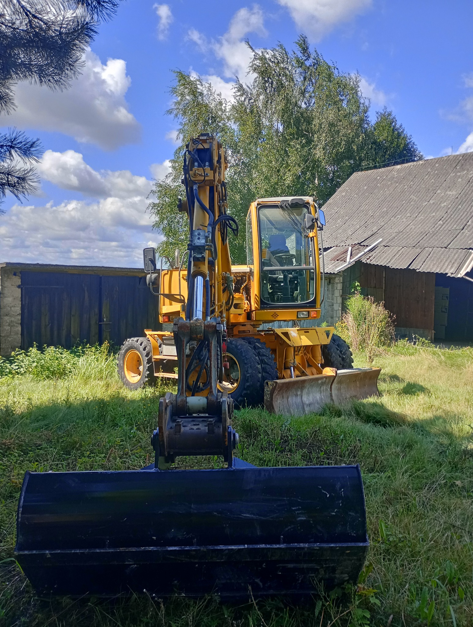 Żółta koparka kołowa stoi na trawiastym terenie, z czarną łyżką skierowaną w stronę obiektywu, na tle budynku gospodarczego i błękitnego nieba z chmurami.