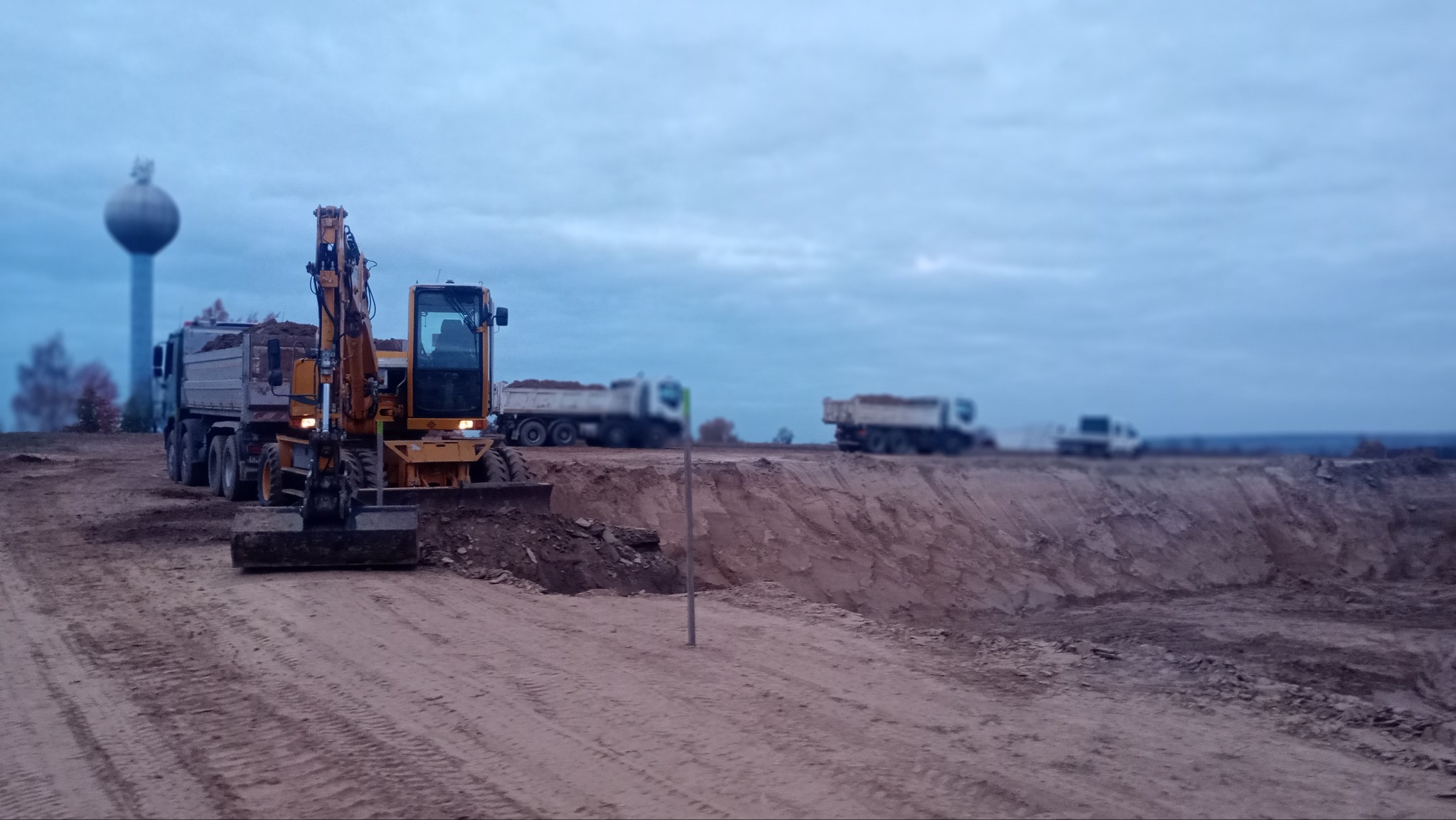 Żółta koparka i ciężarówka z ziemią na tle wykopu, w oddali widoczne inne ciężarówki i wieża ciśnień w pochmurny dzień.