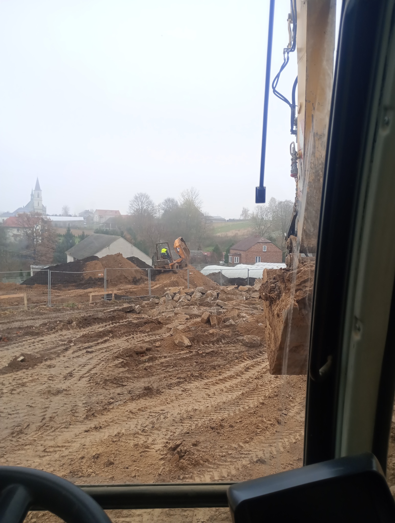 Widok z kabiny koparki na plac budowy z gruzem i koparką w tle, w oddali zabudowania wiejskie i kościół w mglisty dzień.