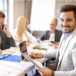 Tomasz Klein Ekspert Finansowy Credipass Polska S.A. oddział Gdynia - Zespół finansowy podczas spotkania: mężczyzna w jasnej marynarce uśmiecha się, pracując na laptopie, w tle współpracownicy analizują dokumenty i rozmawiają przez telefon.