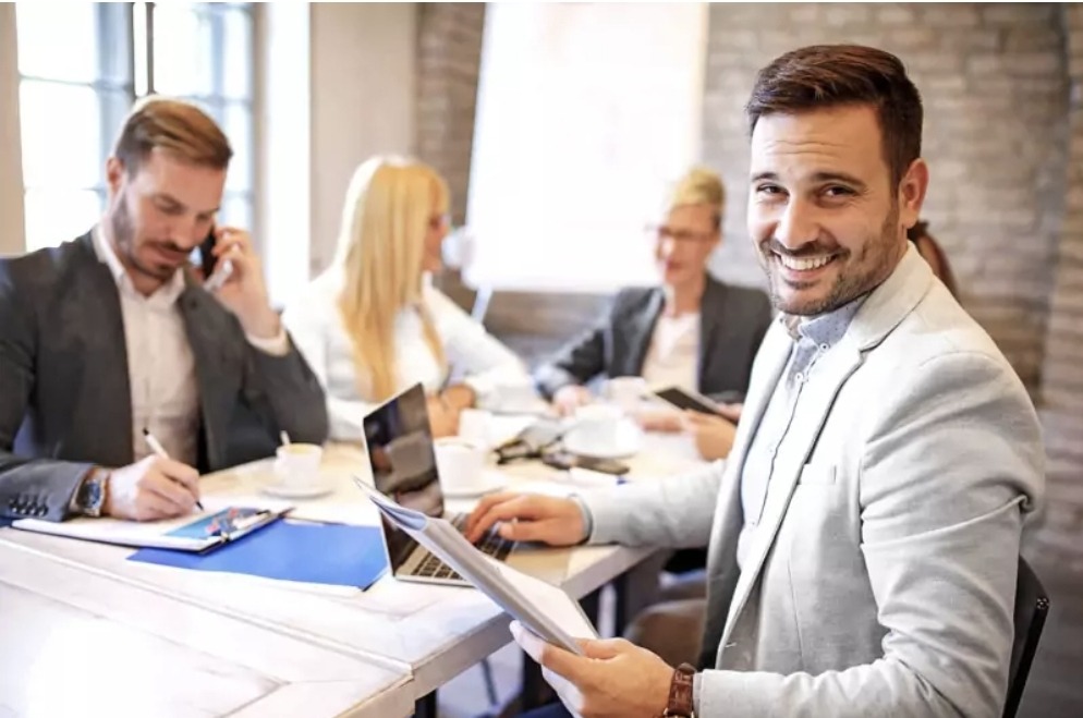Zespół finansowy podczas spotkania: mężczyzna w jasnej marynarce uśmiecha się, pracując na laptopie, w tle współpracownicy analizują dokumenty i rozmawiają przez telefon.