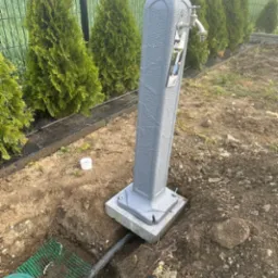 Szara, zewnętrzna kolumna z kranem zamontowana na betonowej podstawie, z widocznymi przyłączami wodnymi w wykopie ziemnym i szeregiem tuj w tle.