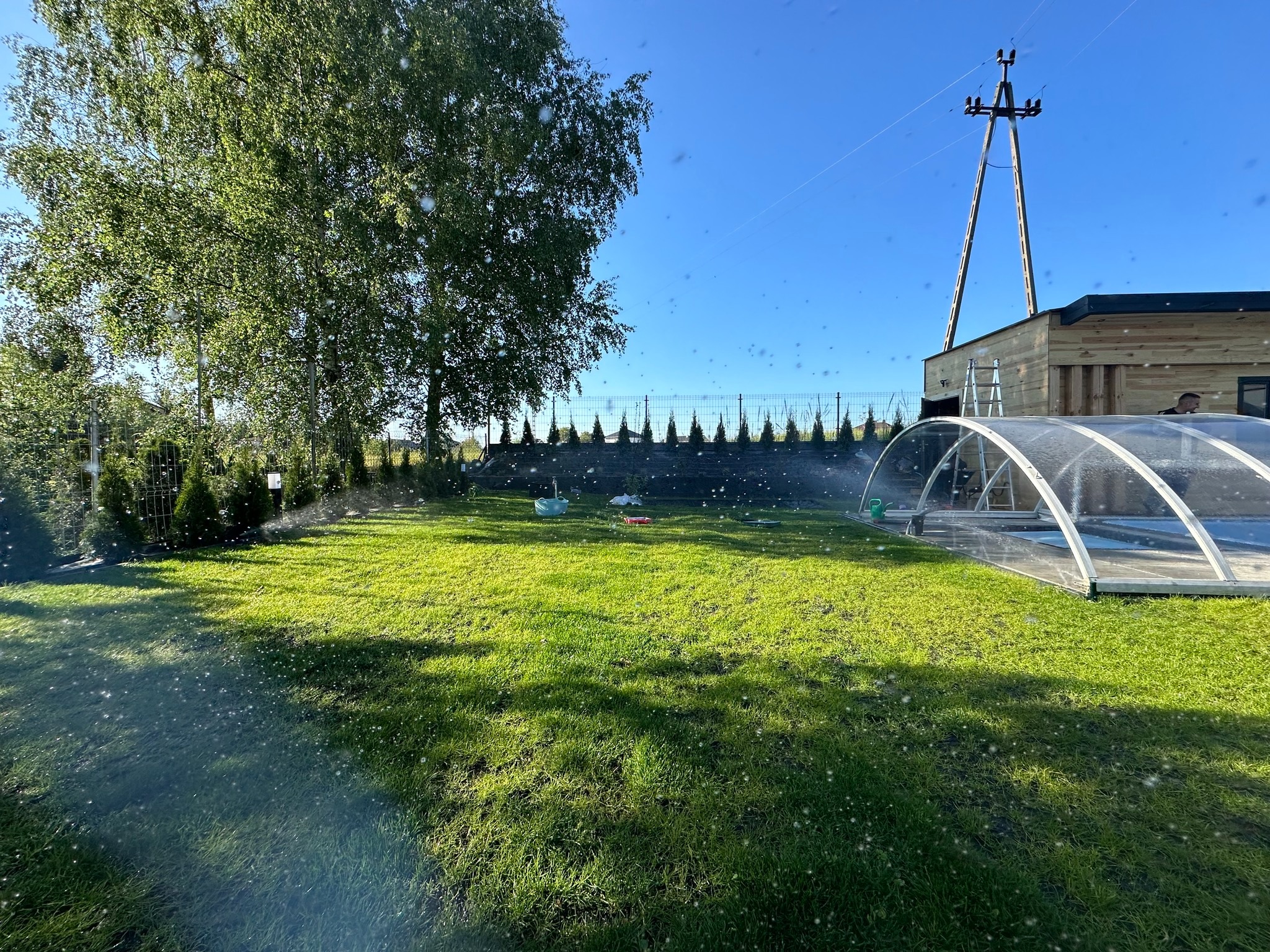 Intensywnie zielony trawnik w trakcie nawadniania przez zraszacze, widoczny basen z zadaszeniem, drzewo liściaste i słup wysokiego napięcia w tle.