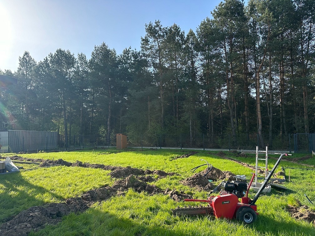 Przygotowanie terenu pod ogród: widok na wykopane rowy w trawie, czerwona glebogryzarka w pierwszym planie, w tle wysokie drzewa i ogrodzenie.